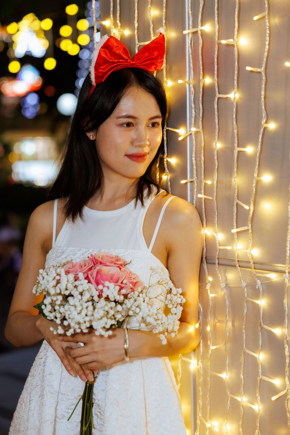 Person in white dress holding pink roses and baby's breath against backdrop of hanging fairy lights.