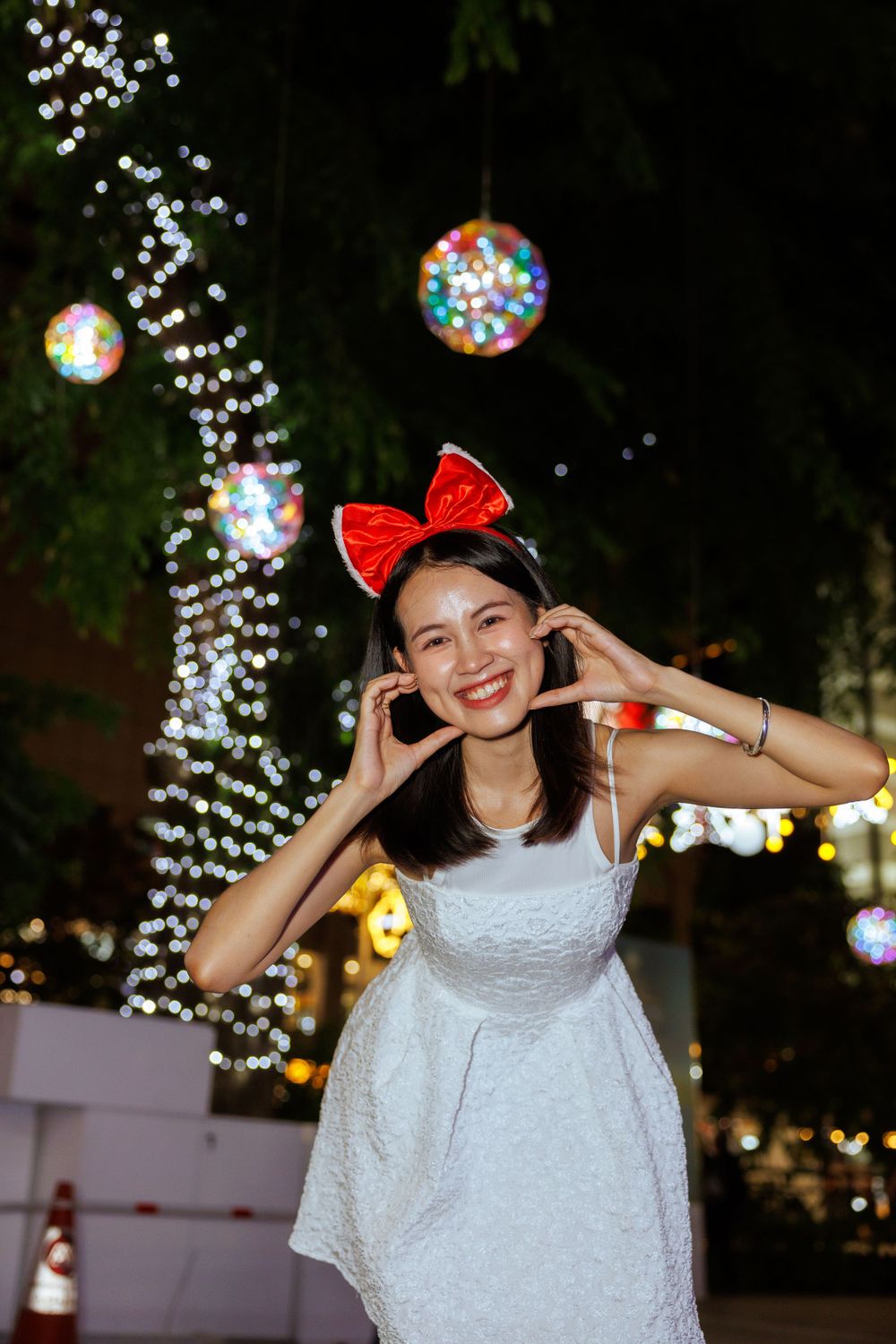 Person in white dress and red bow making playful gesture against backdrop of holiday lights.