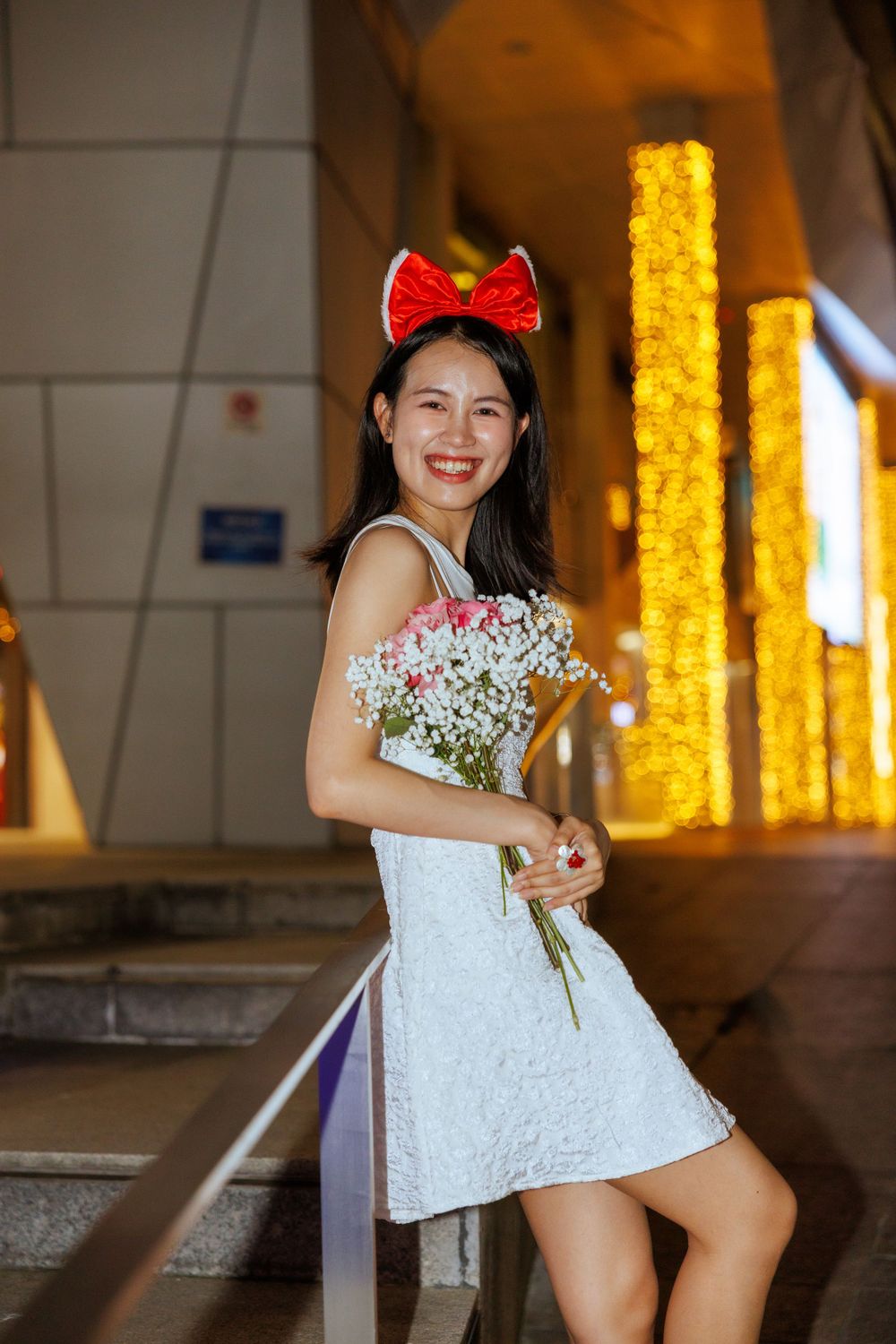 Person in white dress holding flowers poses near illuminated gold pillars with red bow accessory.