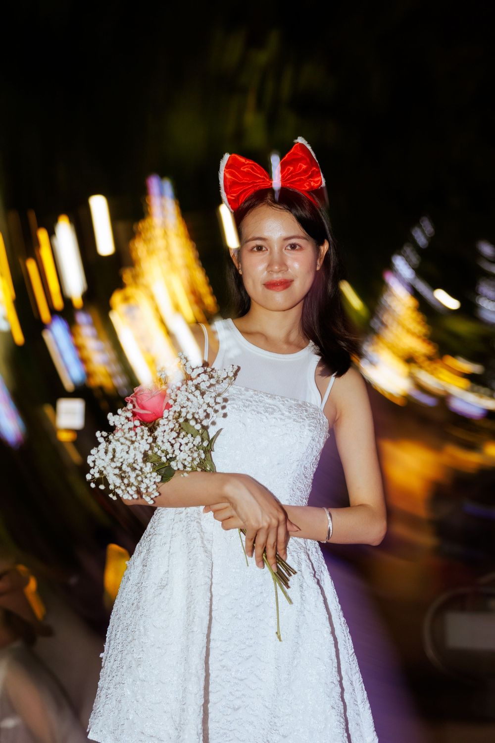 A person in a white dress holds baby's breath flowers against a blurred cityscape at night.