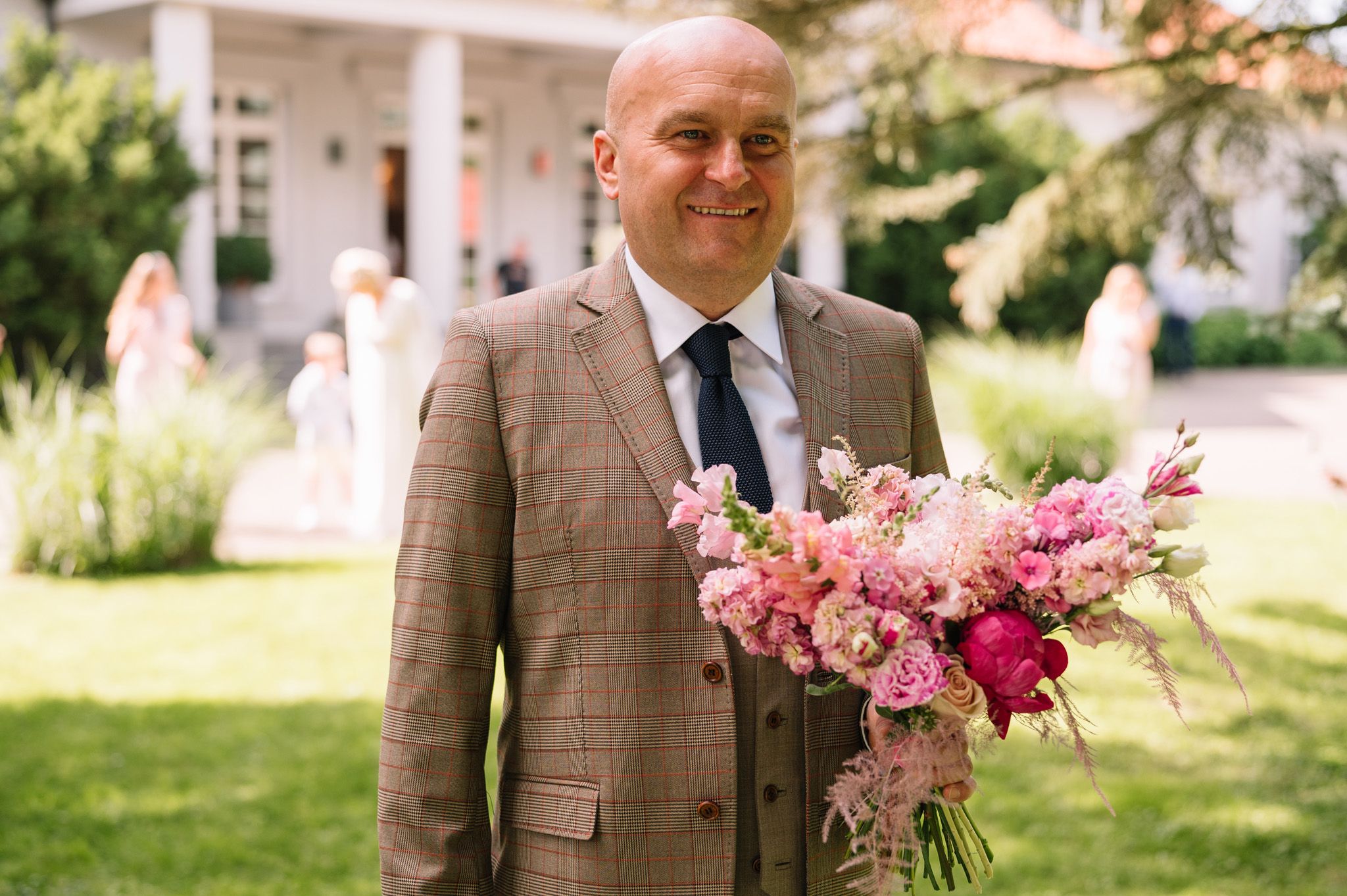 Someone in a brown suit holds a vibrant pink and magenta floral bouquet at an outdoor wedding.