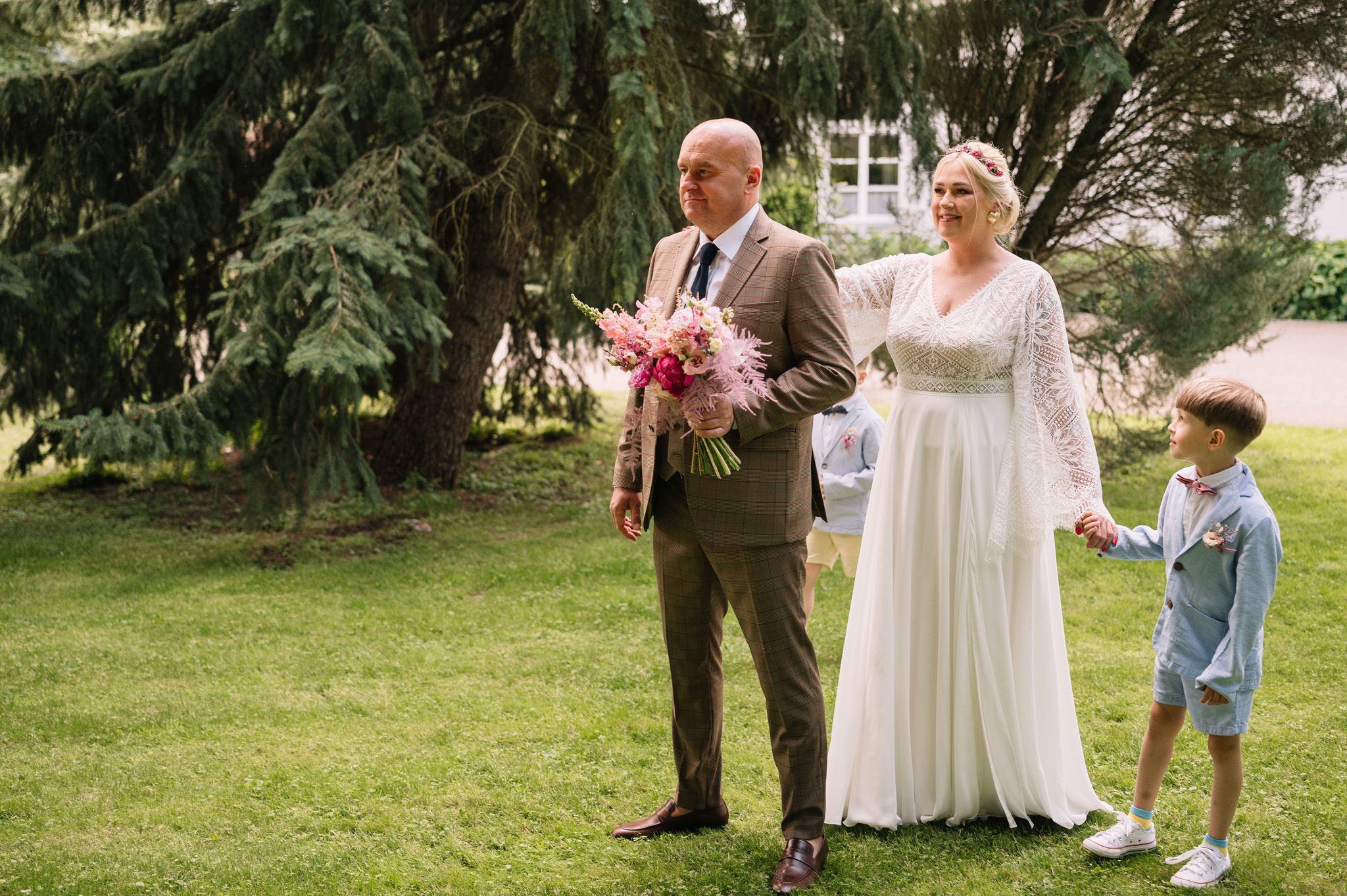 A couple stands on a lush lawn holding pink flowers during their wedding celebration.