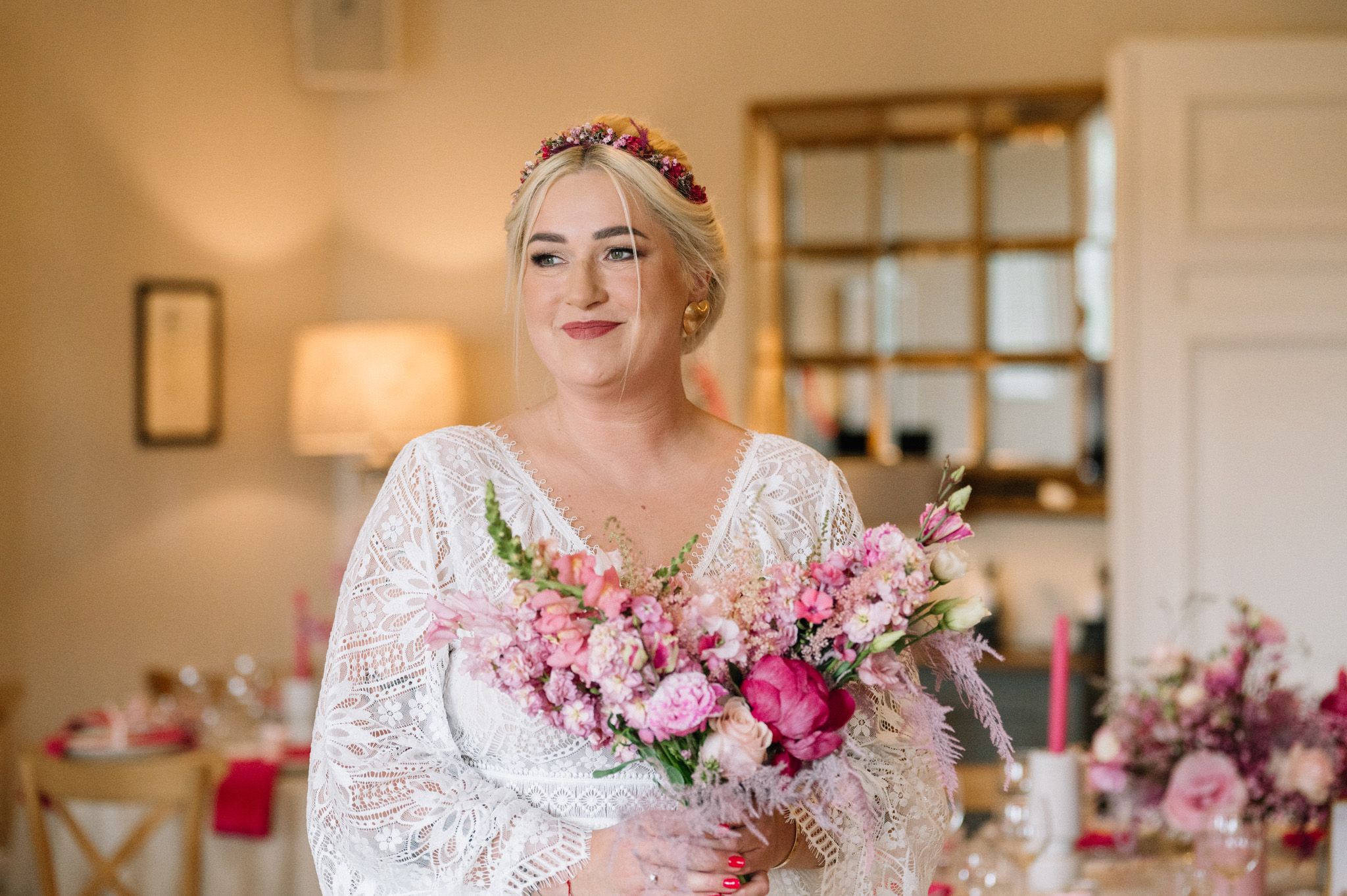 A bridal portrait featuring a lace dress and pink floral bouquet in a warmly lit room.