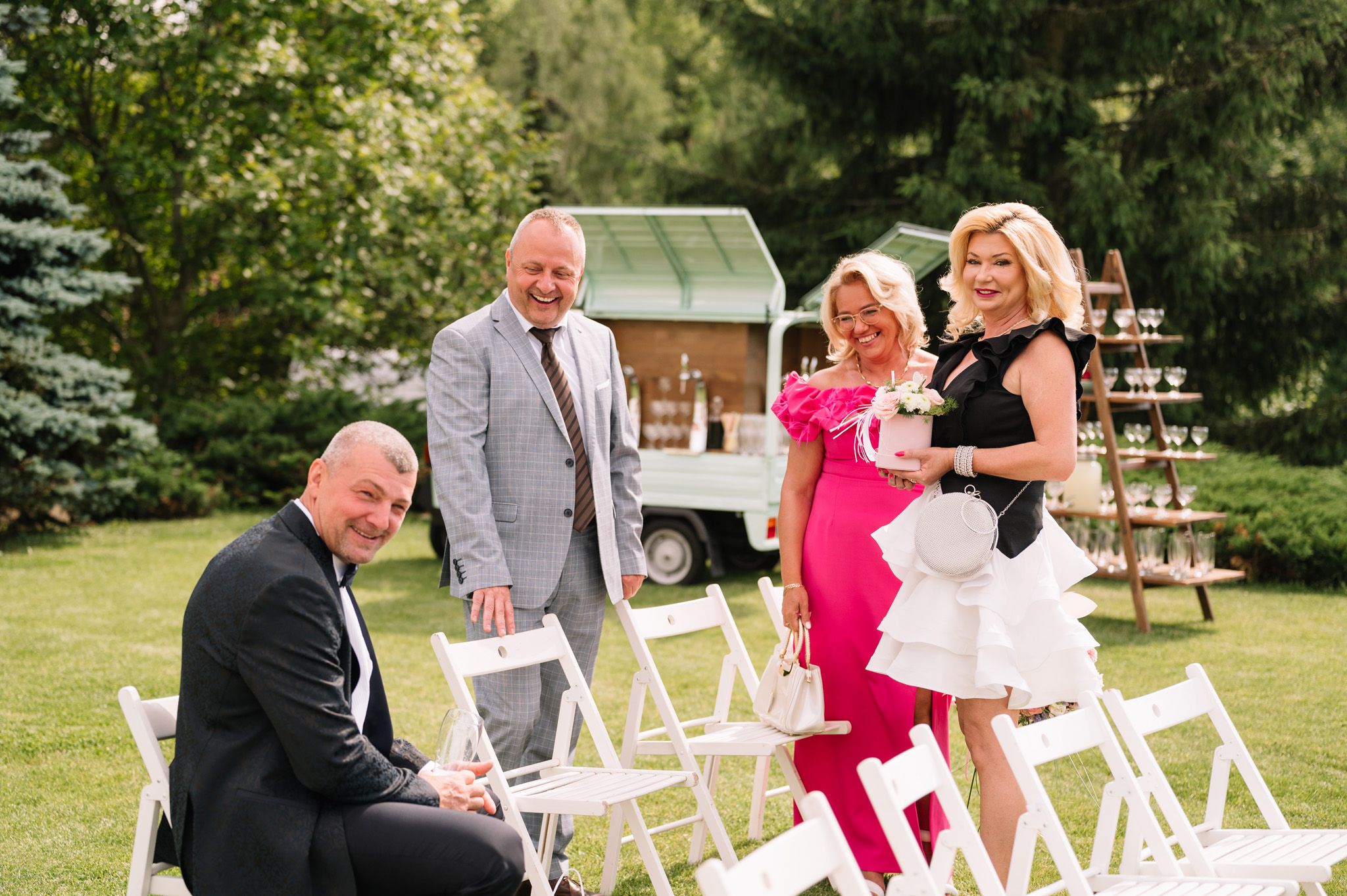 Wedding guests gather around white chairs and a champagne tower setup in an outdoor garden setting.