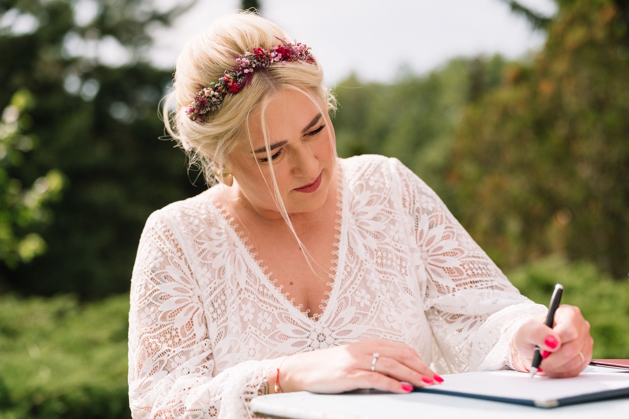 Someone in a lace white dress wearing a floral crown writes in a journal outdoors.