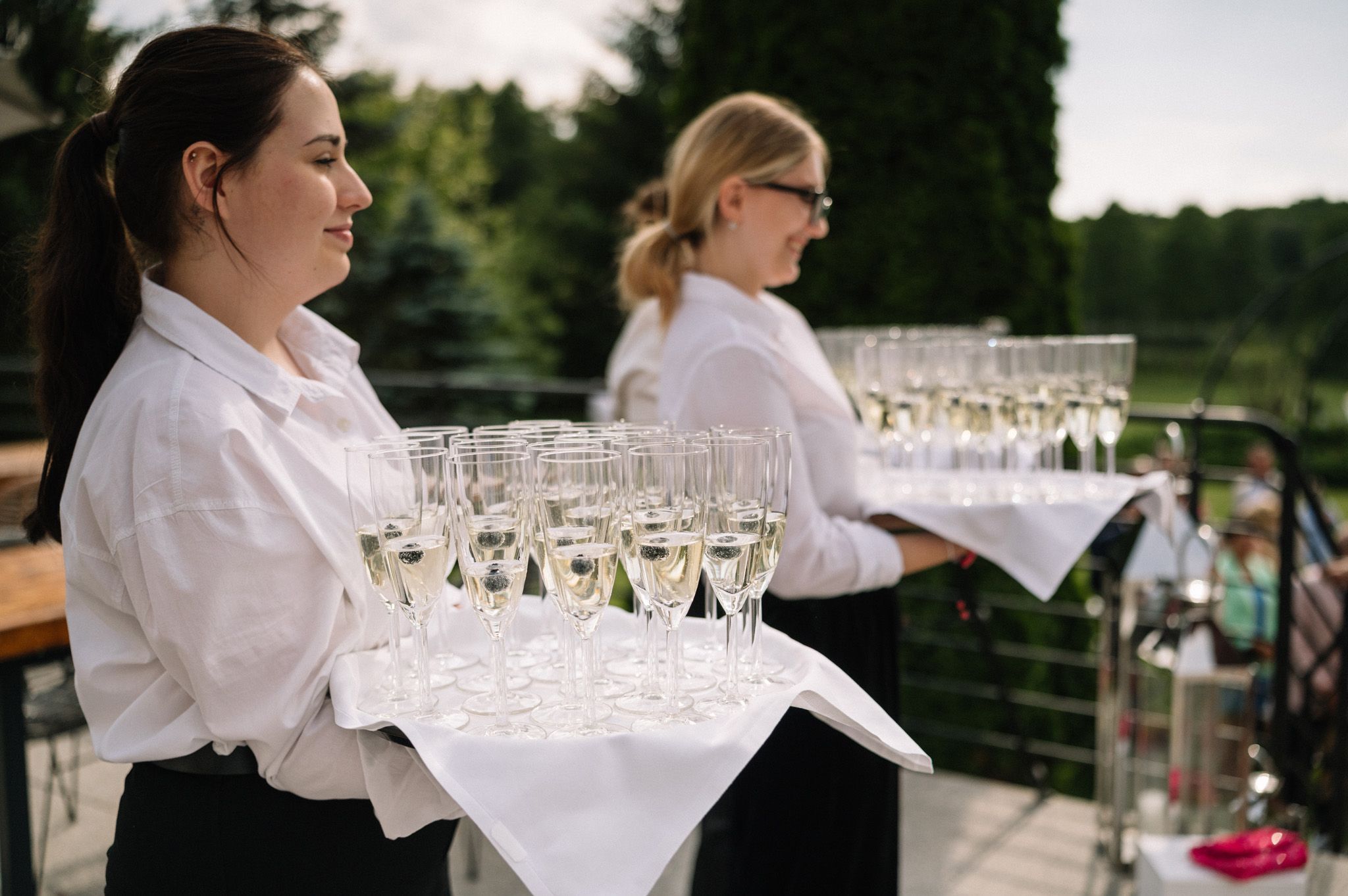 Servers in white shirts holding trays of champagne glasses at an outdoor event.