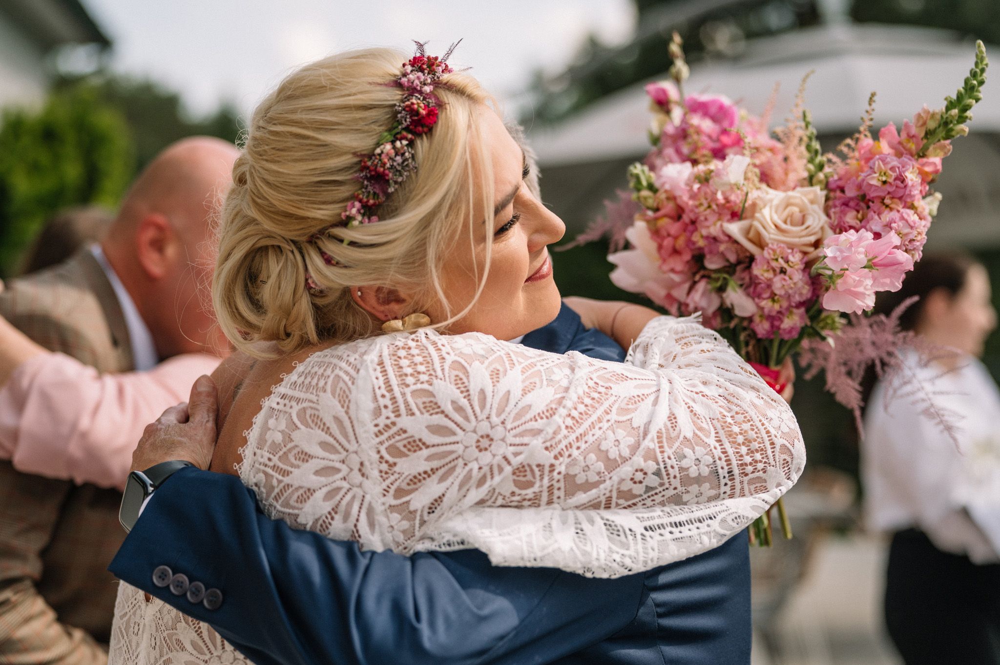 An emotional embrace featuring lace details and pink floral bouquet at a wedding celebration.