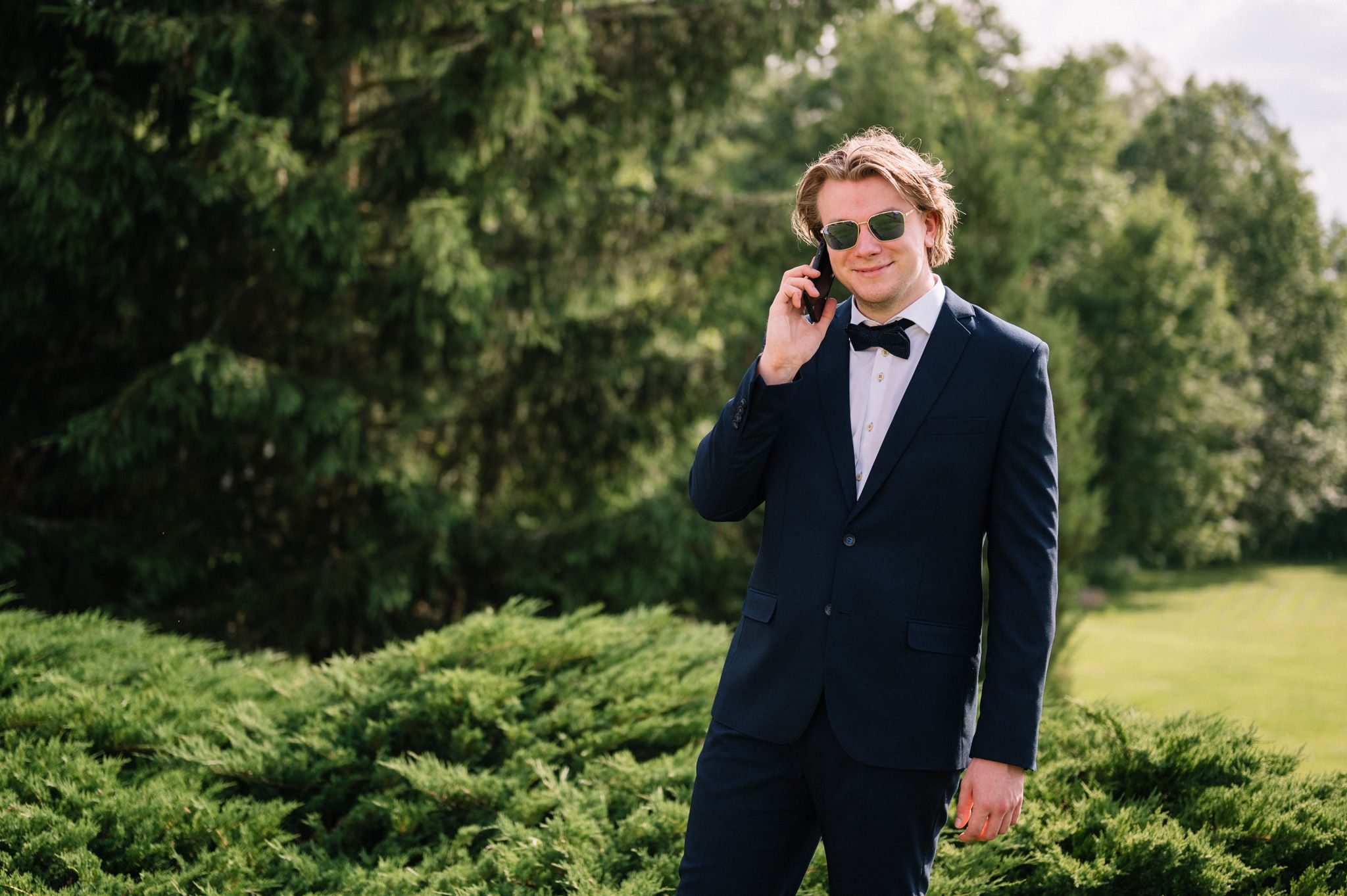 Formal-dressed guest in navy suit and bowtie makes phone call in garden setting.
