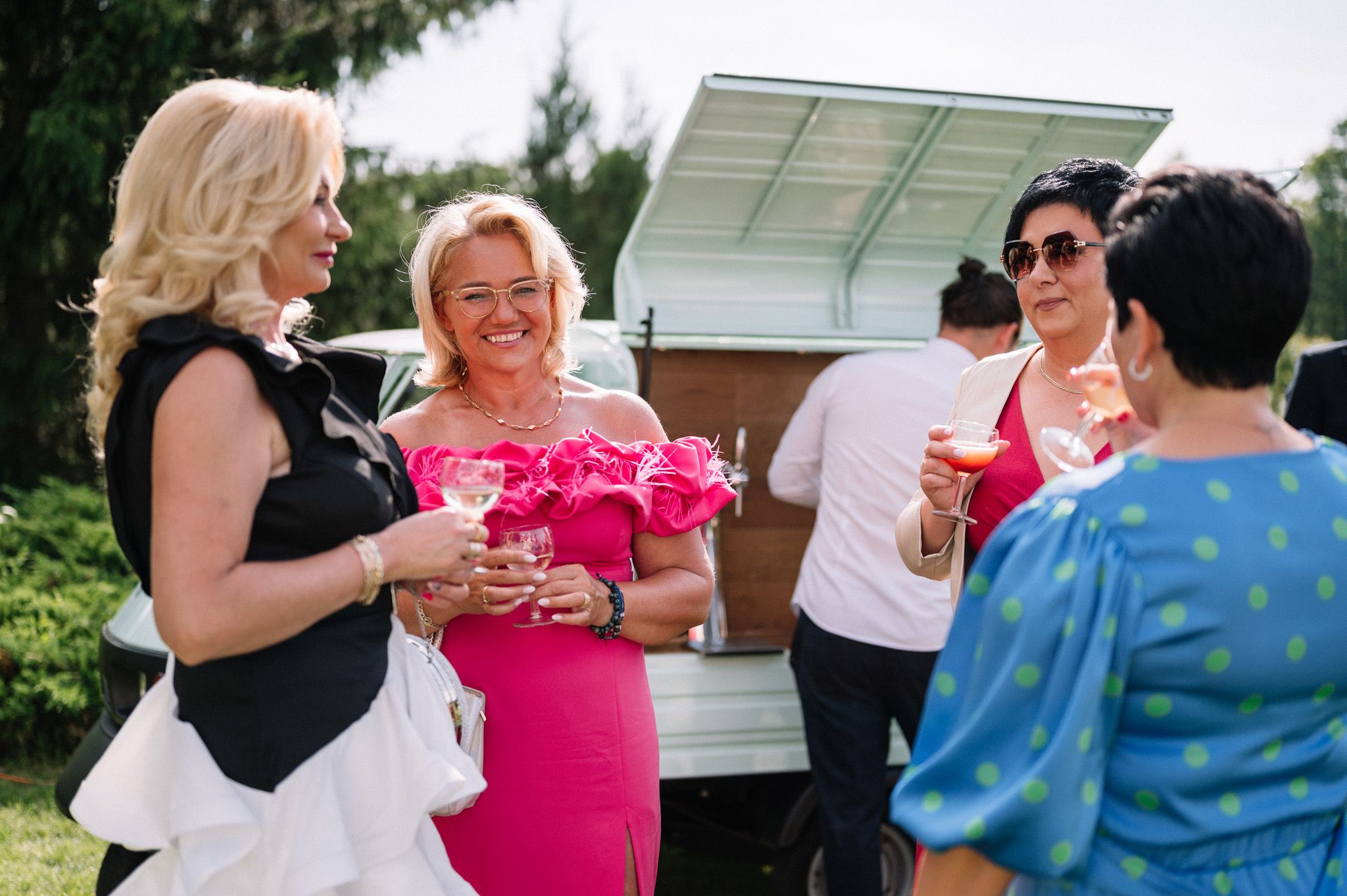 Group of guests sharing drinks and laughter near food truck at outdoor event.
