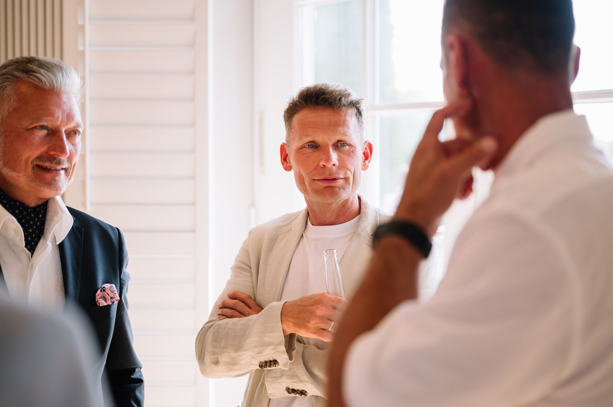 Casual gathering of guests in formal attire having a conversation near white shuttered windows.