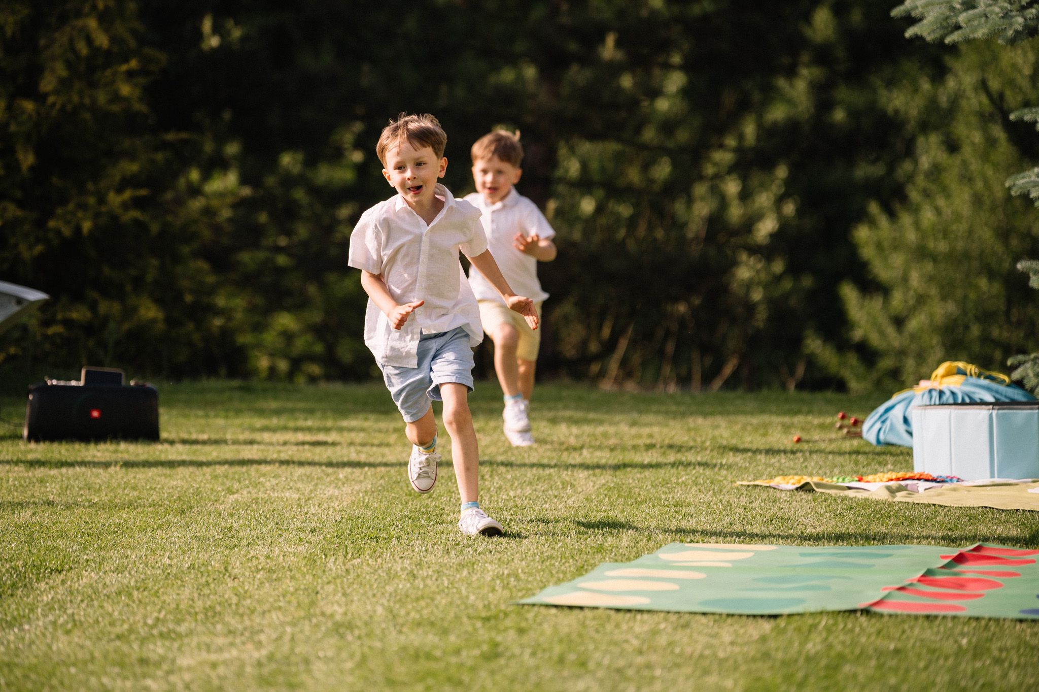 Two children running and playing on a grassy lawn with picnic blankets in the background.