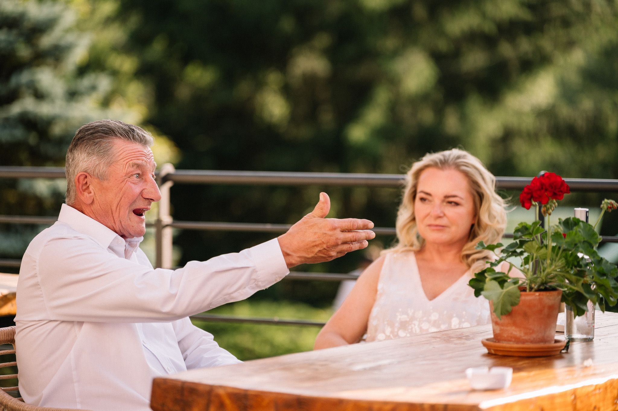 Two people engaged in animated conversation at an outdoor table with potted flowers.