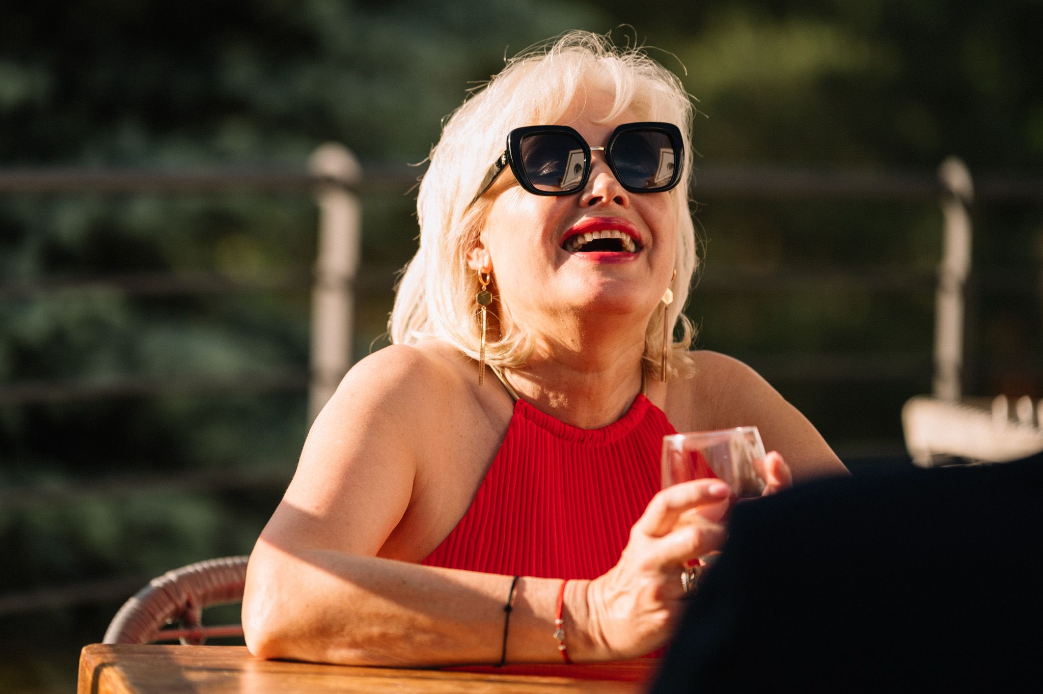 Woman in red top and sunglasses laughing joyfully at an outdoor gathering.