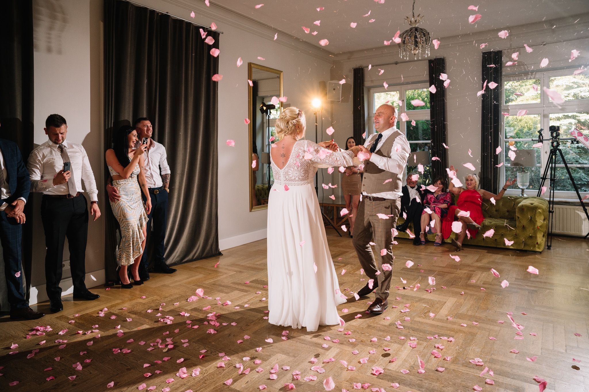 Newlyweds share their first dance as rose petals fall around them in an elegant venue with black curtains.