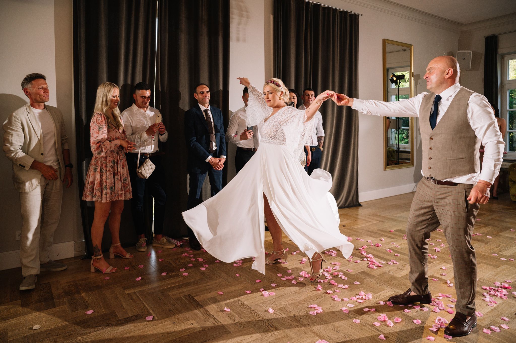 Wedding couple spins and dances while guests watch from the sidelines with rose petals scattered on floor.