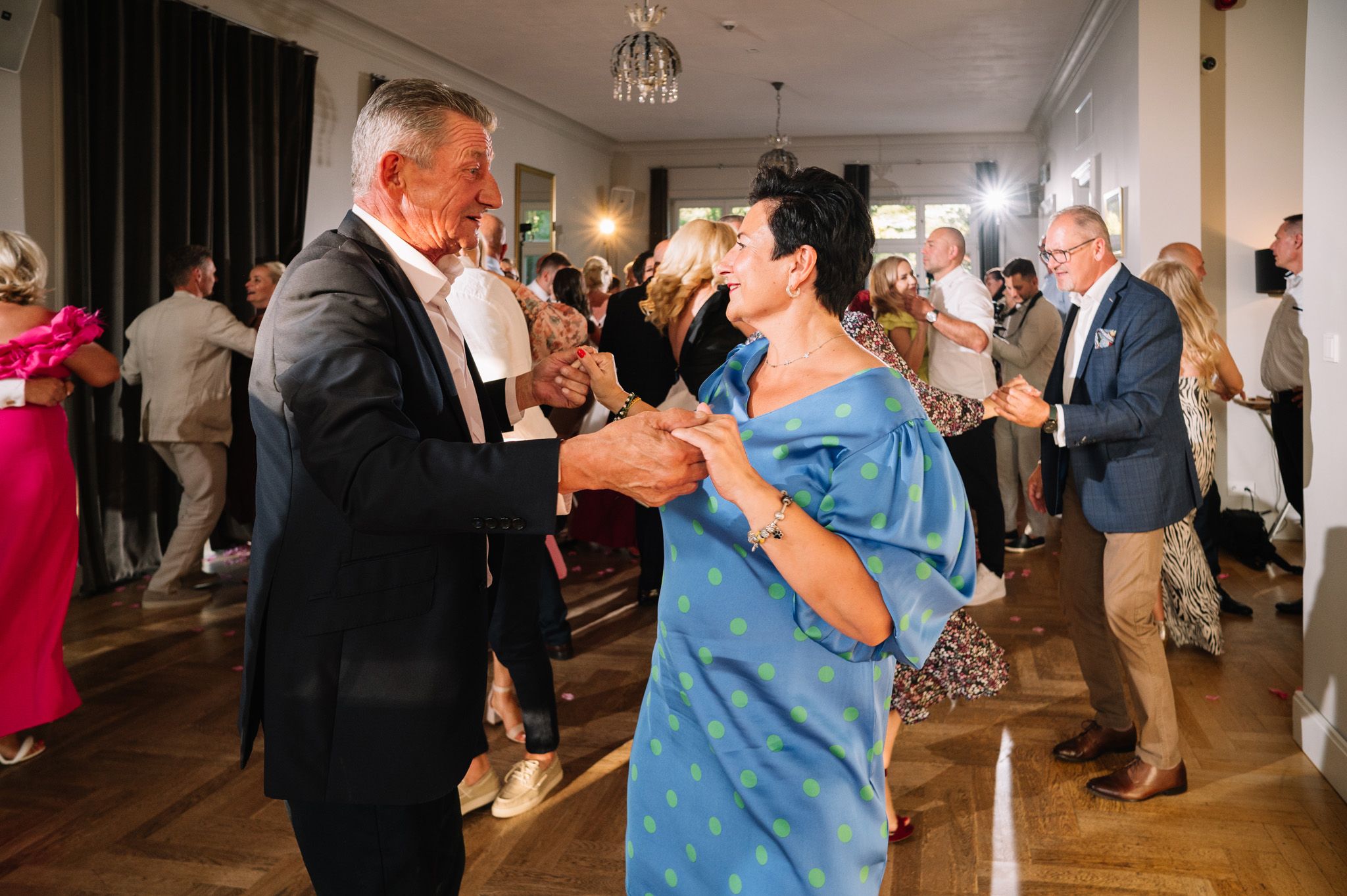 Wedding guests dancing in pairs during an indoor reception with hardwood floors.