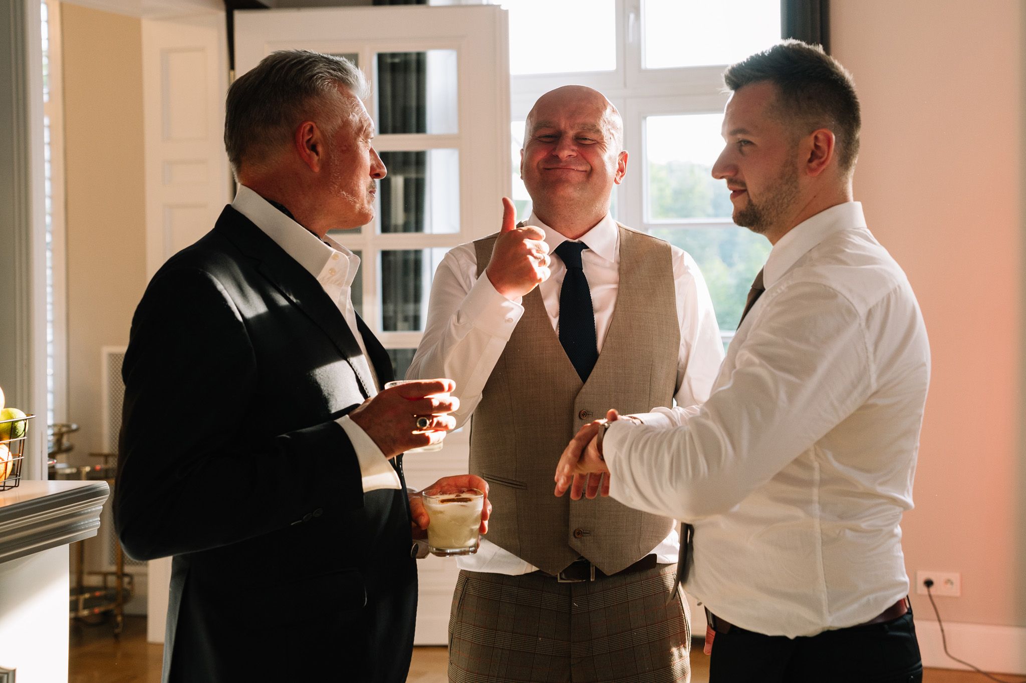 Three men in formal attire having an animated conversation near a bright window.