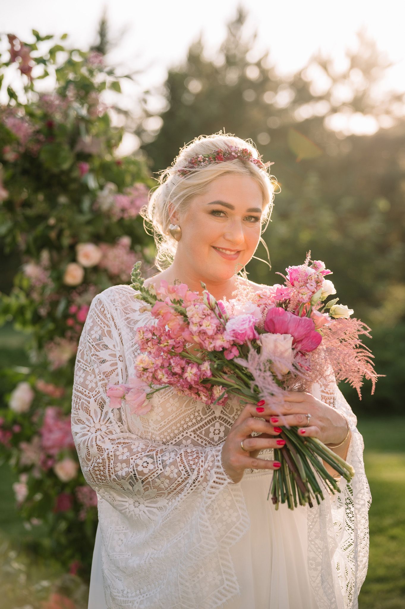 Bride in lace dress holding pink floral bouquet against sunset garden backdrop.