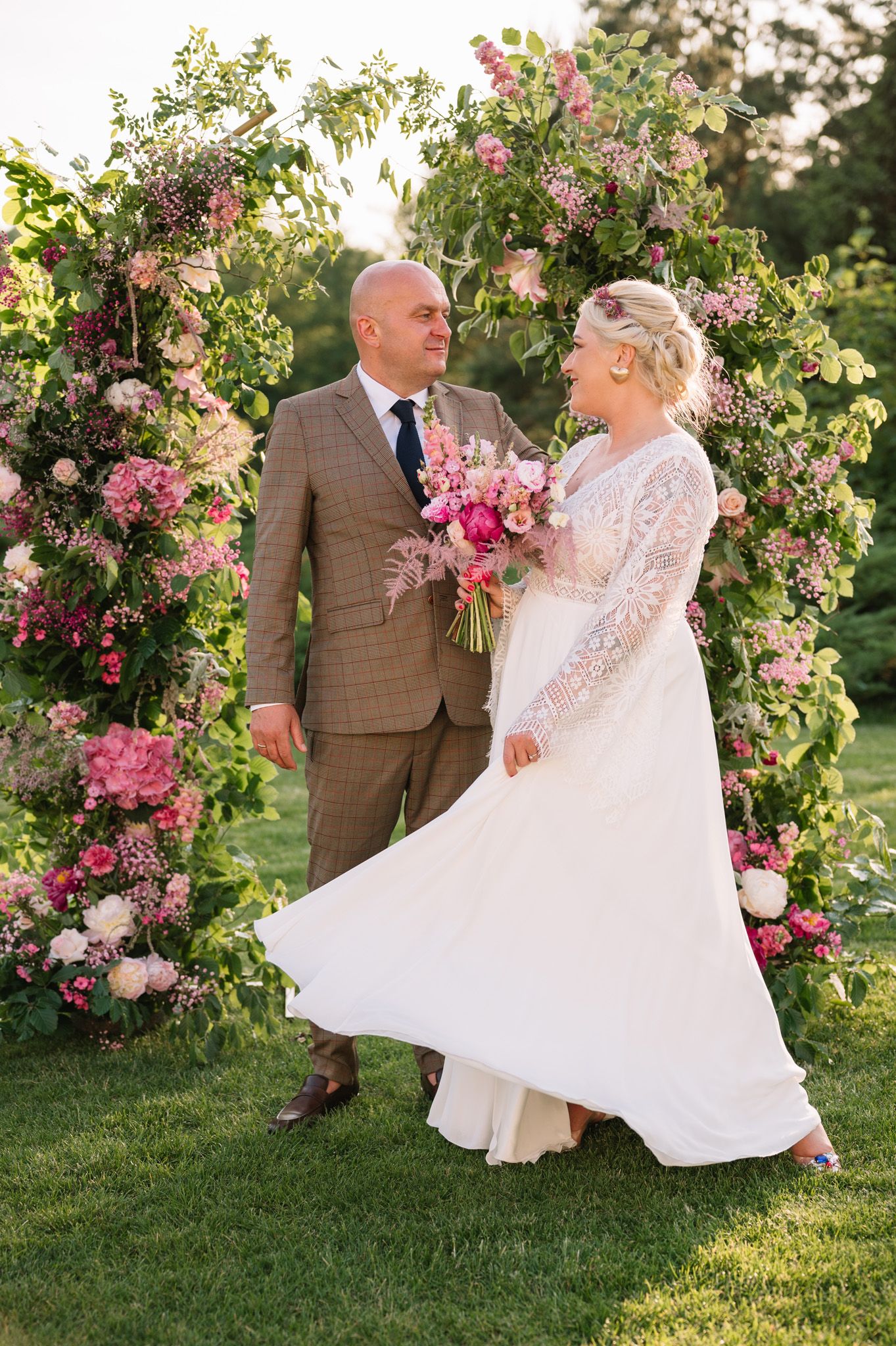 A romantic wedding couple poses under a lush floral arch with pink roses in a garden setting at sunset.