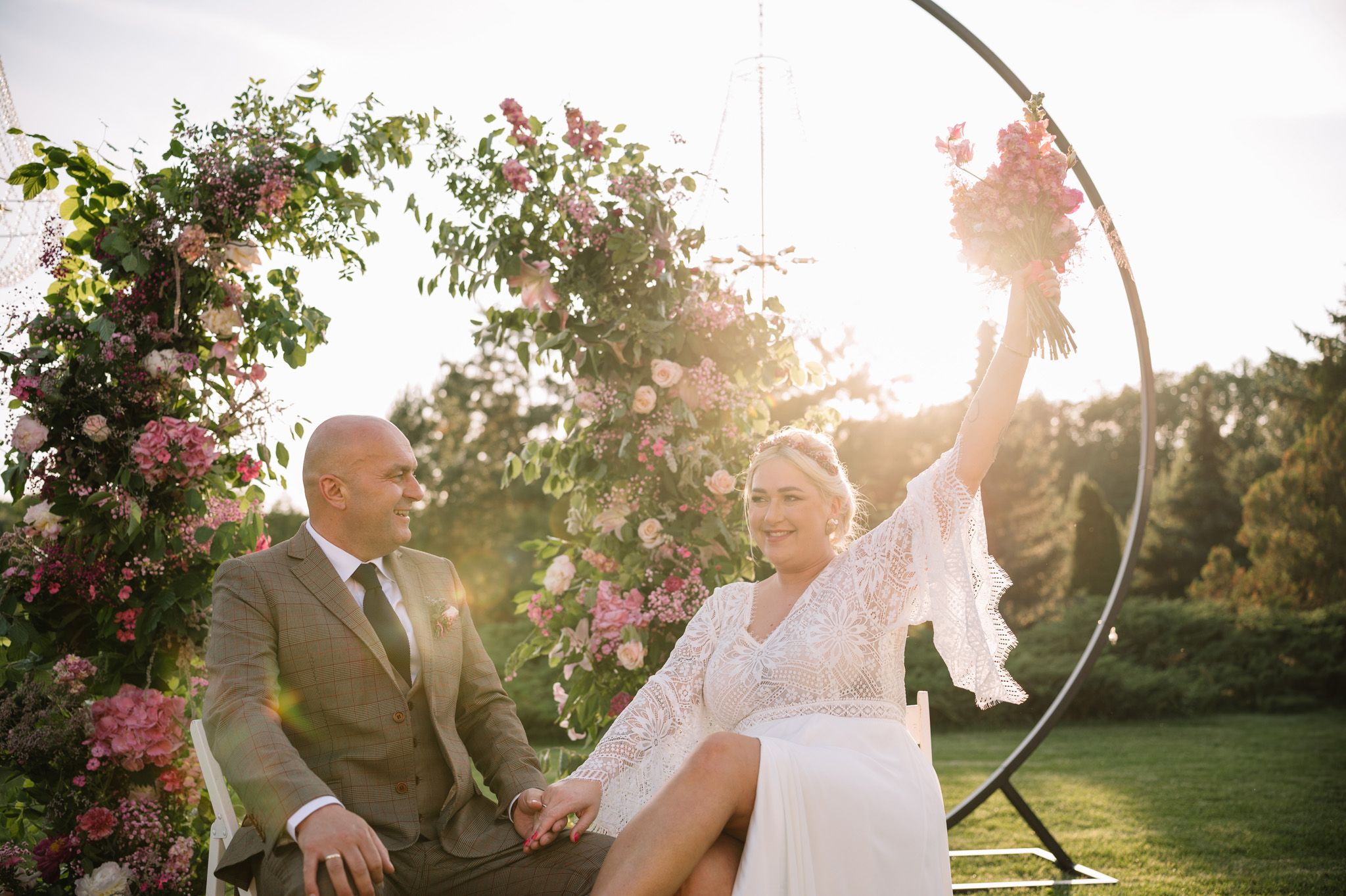Wedding couple shares a playful moment under a floral arch at golden hour.