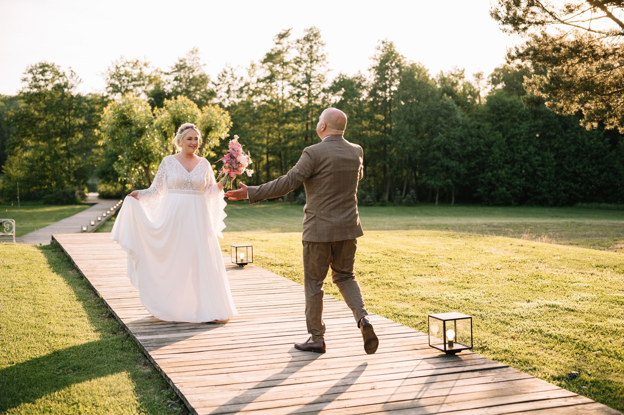 Wedding couple dances joyfully on a wooden boardwalk surrounded by green trees.