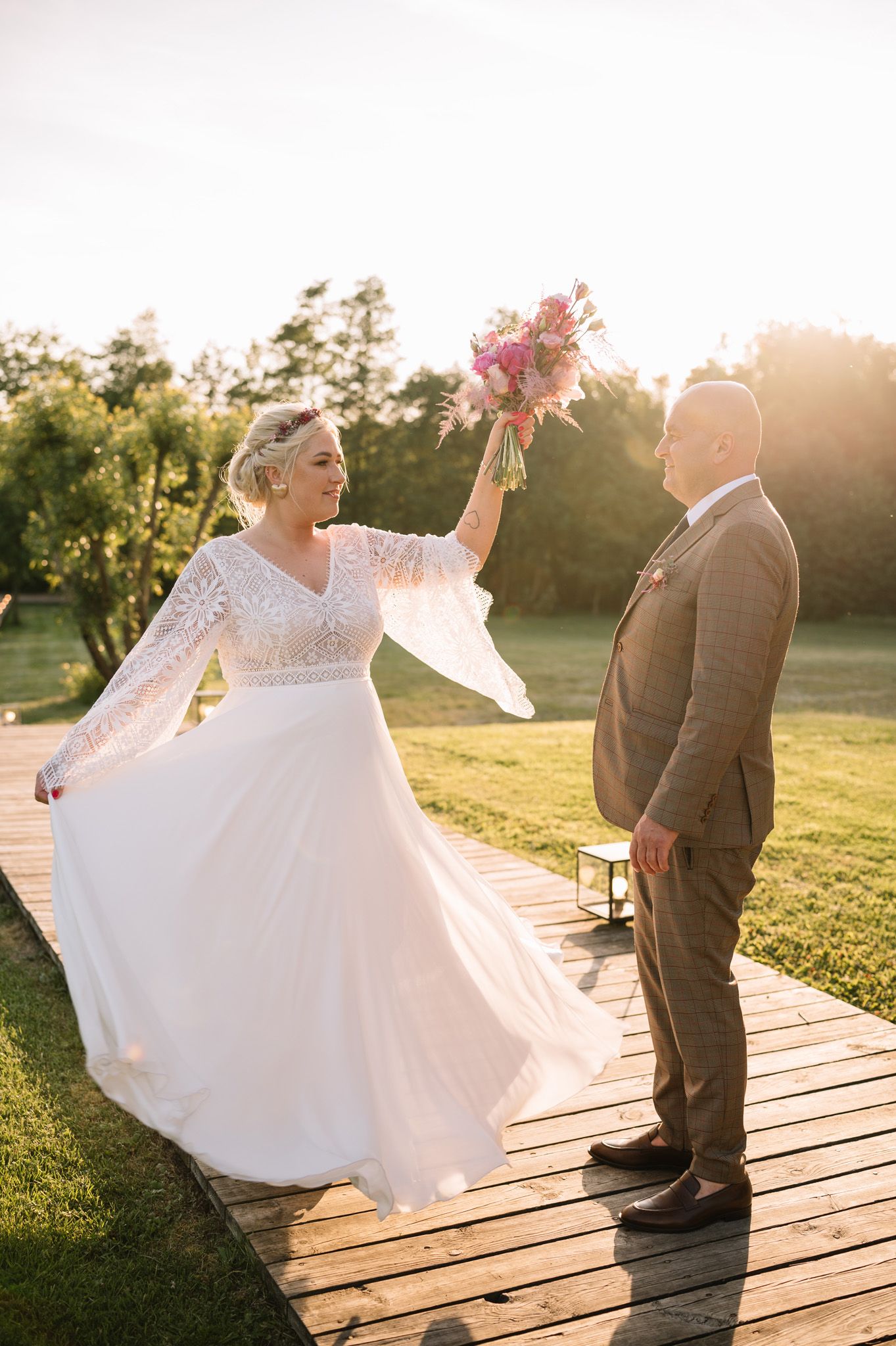 Bride twirls her dress on a wooden deck while holding a pink bouquet at sunset.