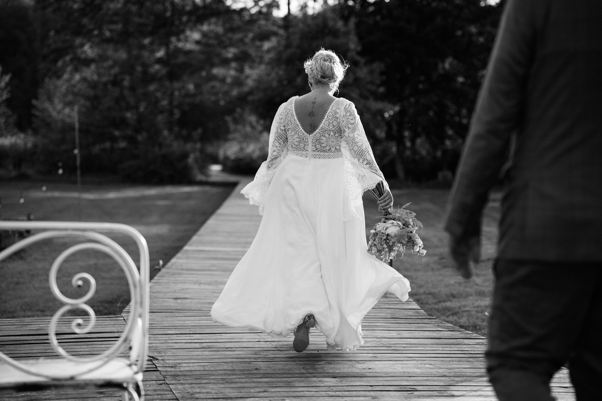 Black and white photo of bride walking along a wooden boardwalk in a flowing lace dress.