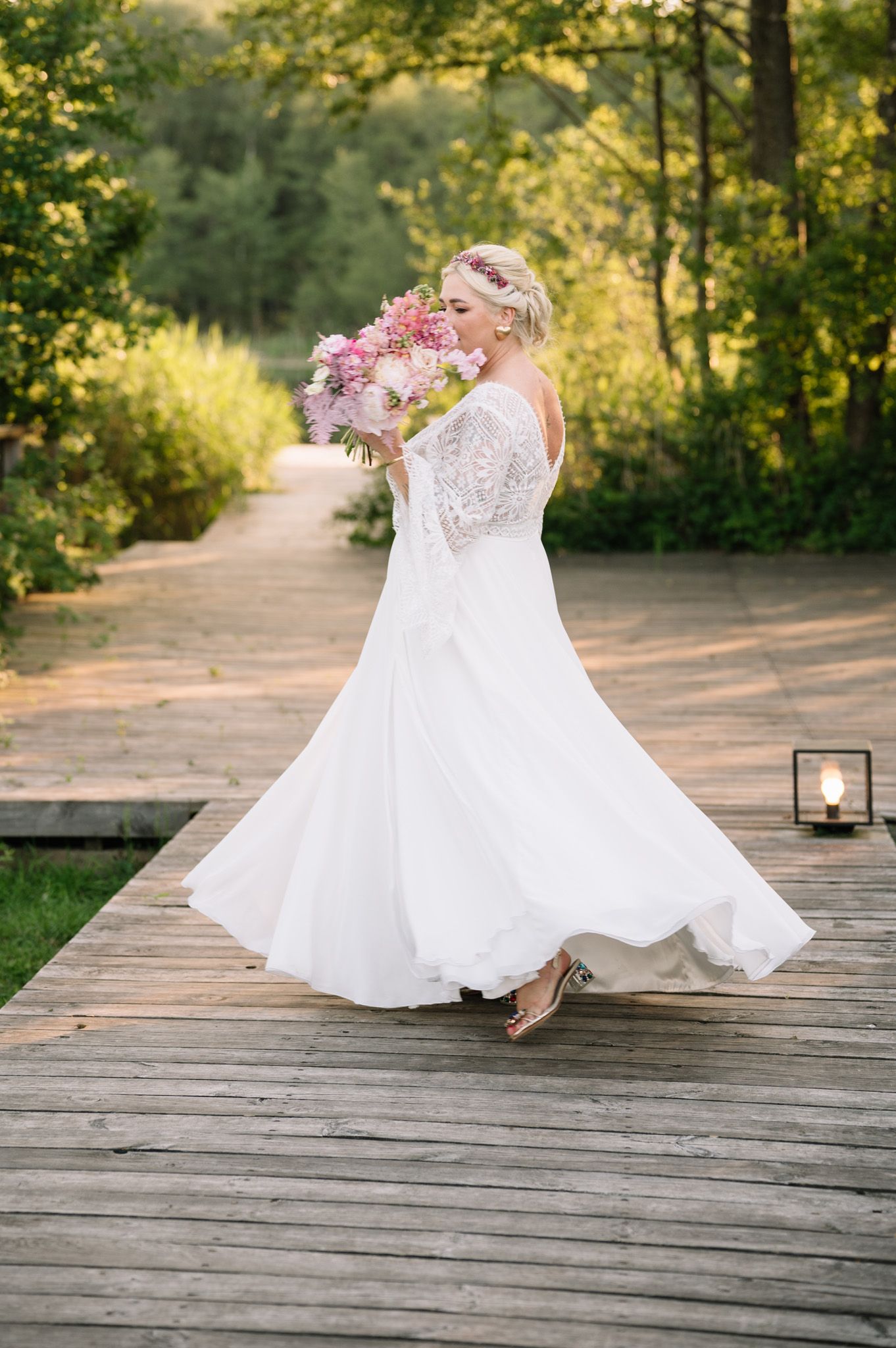 Bride in lace gown twirls with pink bouquet on wooden deck surrounded by summer greenery.
