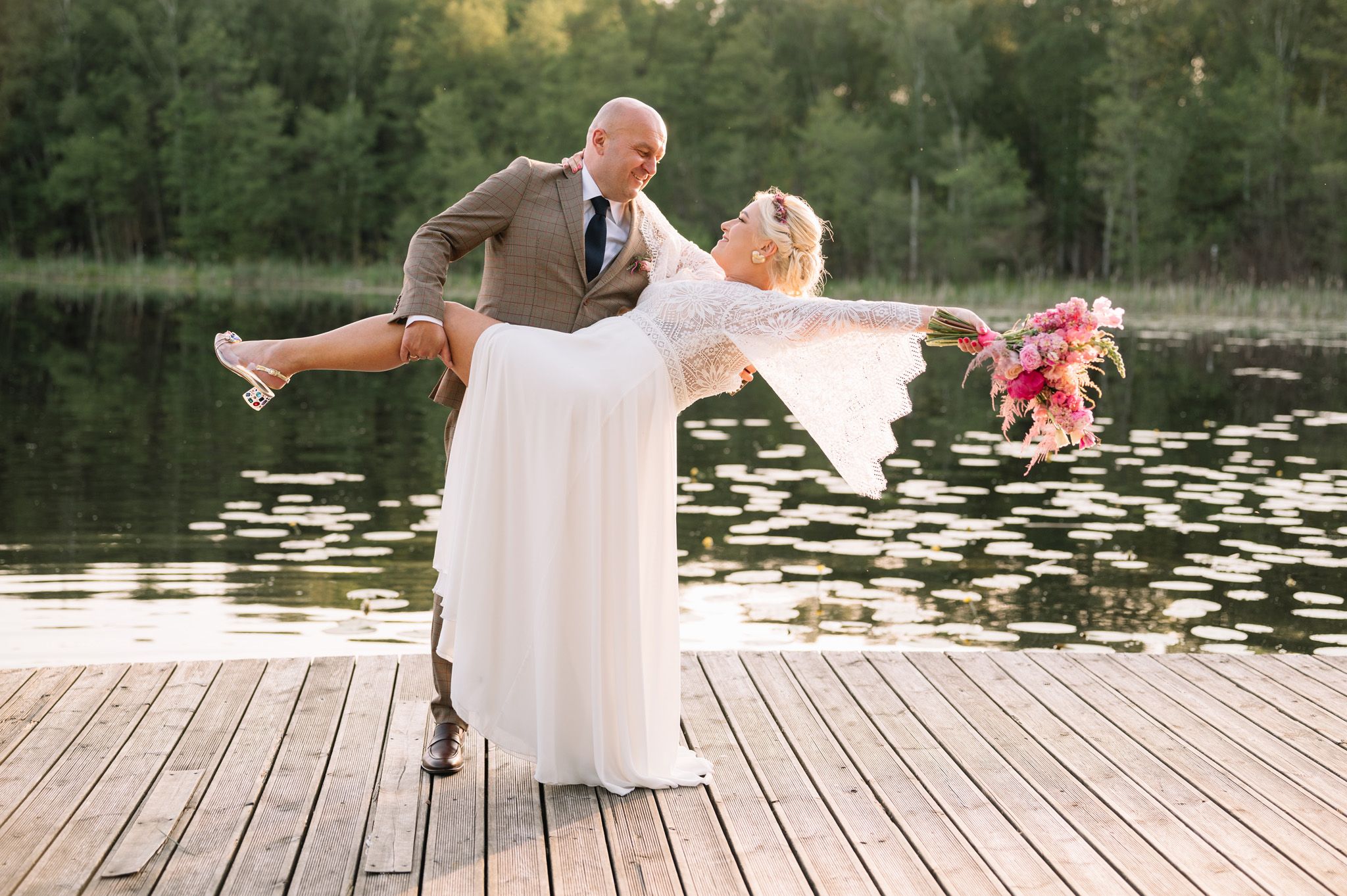 Playful wedding couple strikes dramatic pose on wooden dock by lily pond.