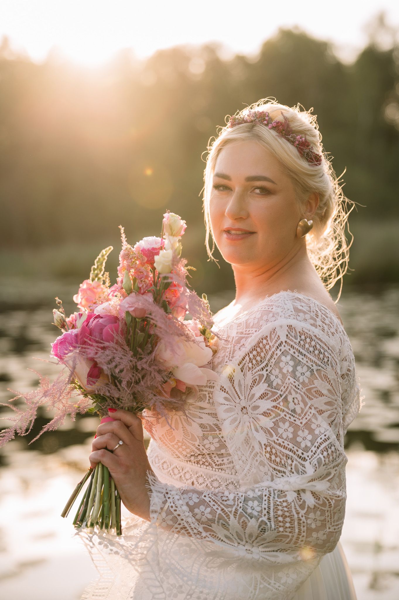 Beautiful lace wedding dress and pink floral bouquet captured in golden sunset light by a lake.