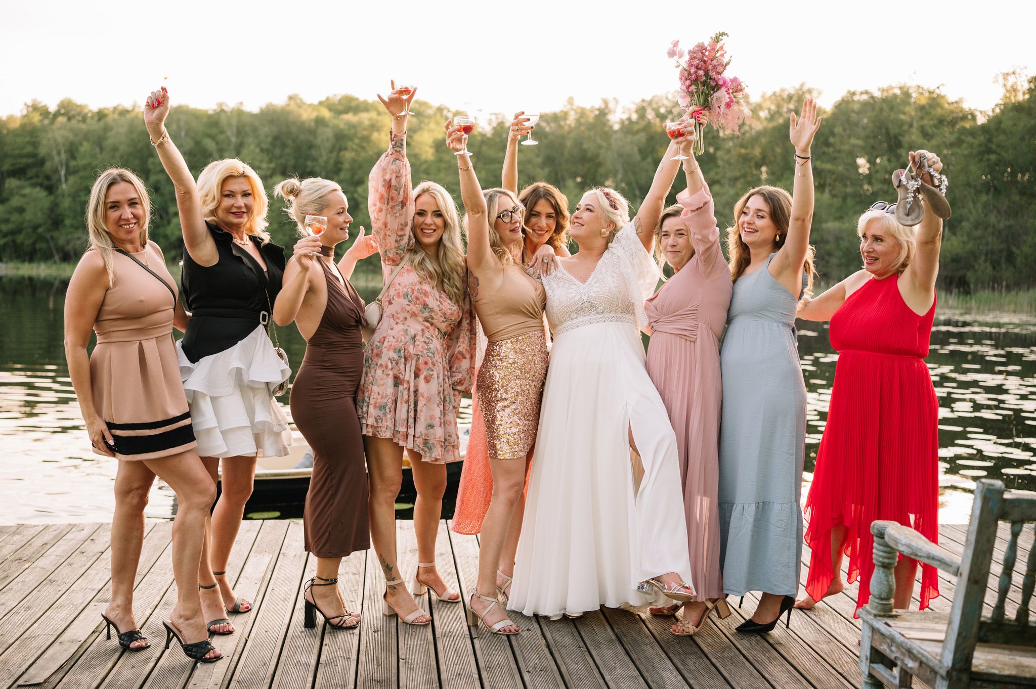 Wedding party of women celebrating on wooden dock by lake at sunset.
