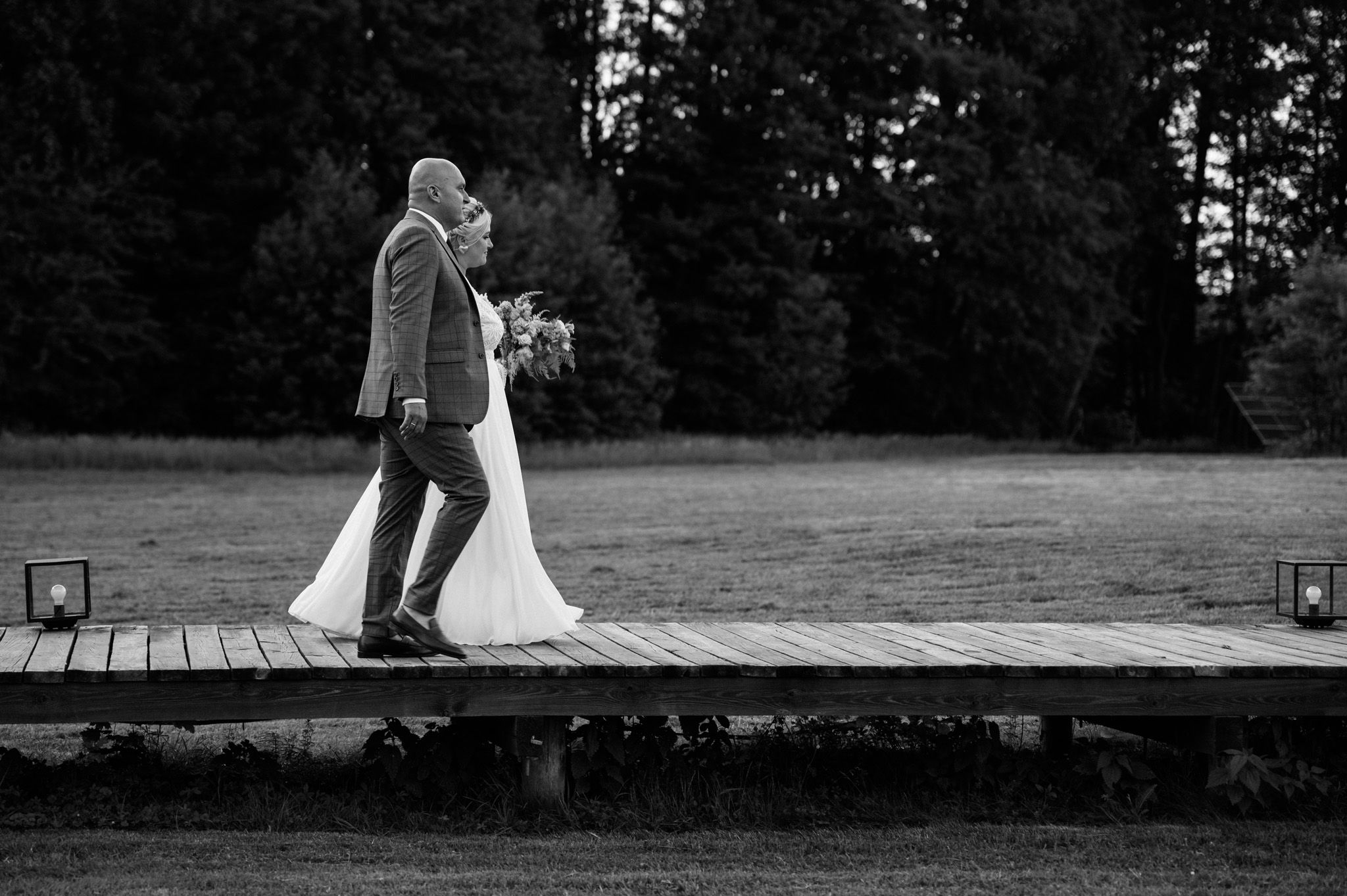 Black and white photo of couple walking on wooden pier by water.
