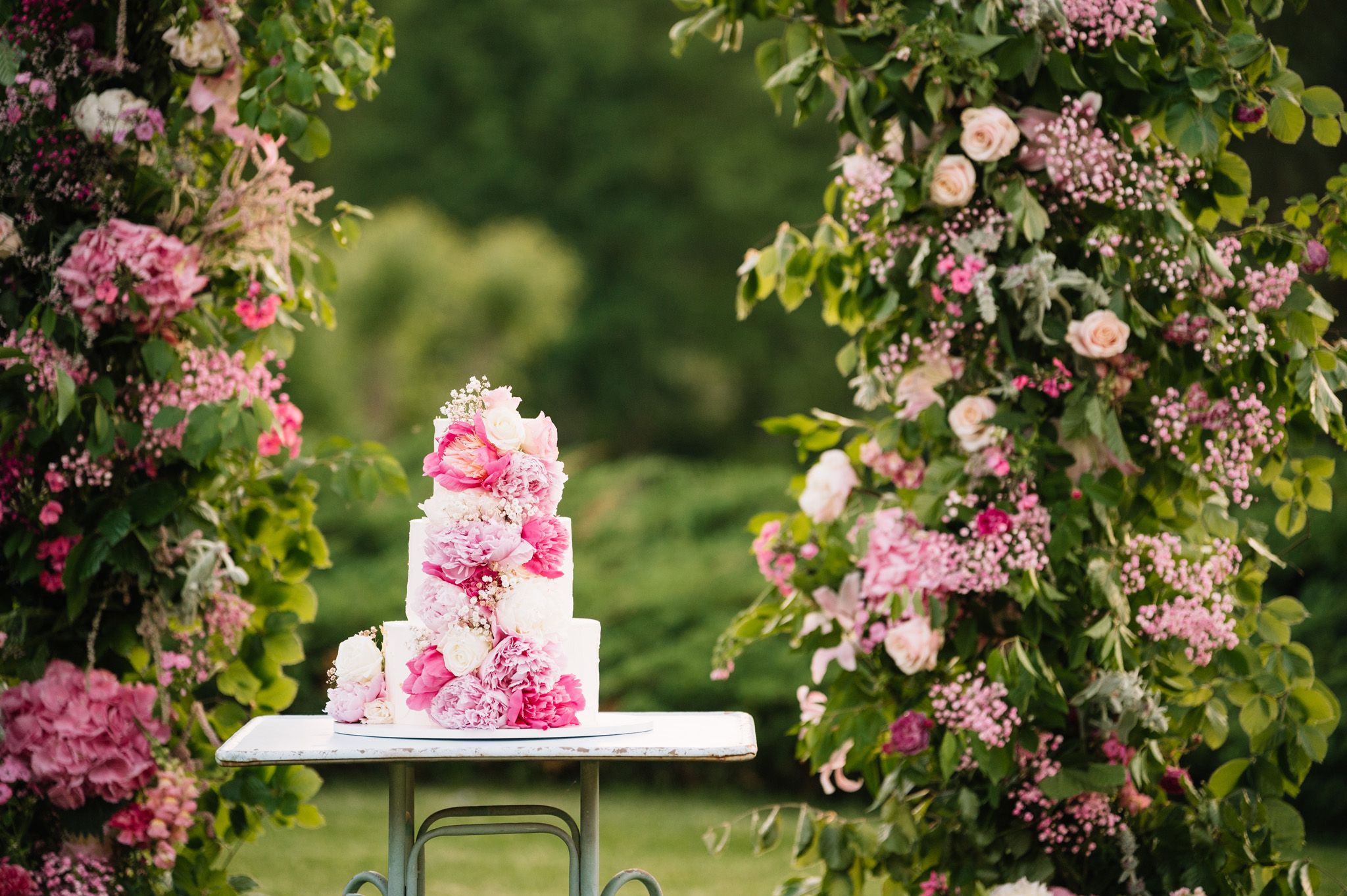 A tiered pink floral wedding cake displayed in a garden setting.