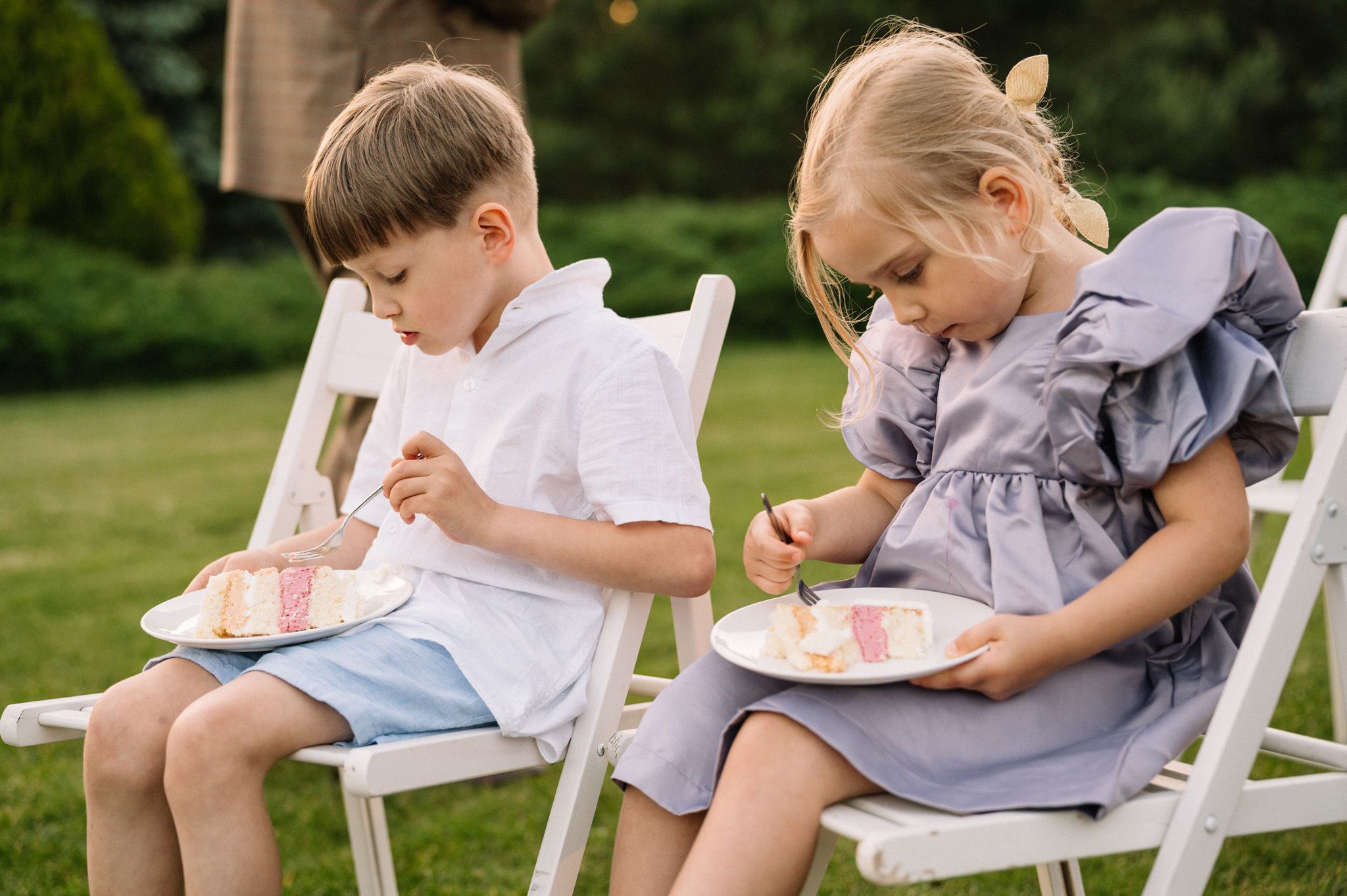 Two children in formal wear eating cake while seated in white chairs at outdoor event.