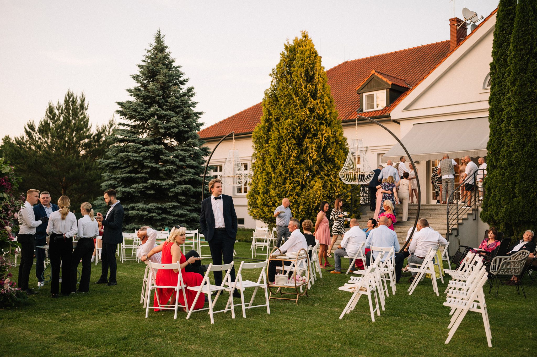Wedding guests gather on a lawn with white chairs in front of a house with red roof and evergreen trees.