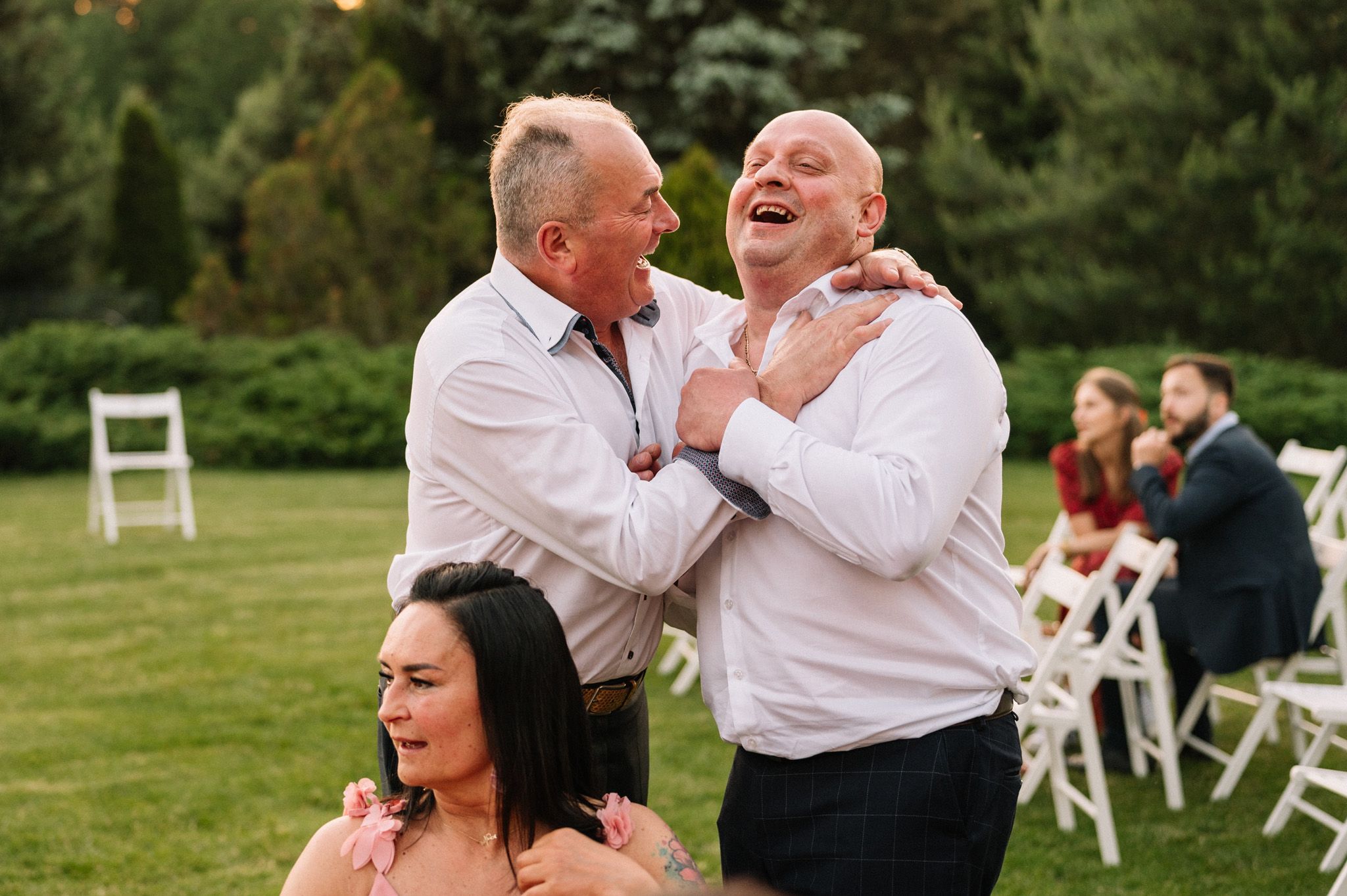 Candid moment of laughter and embraces during outdoor party with white chairs visible in background.