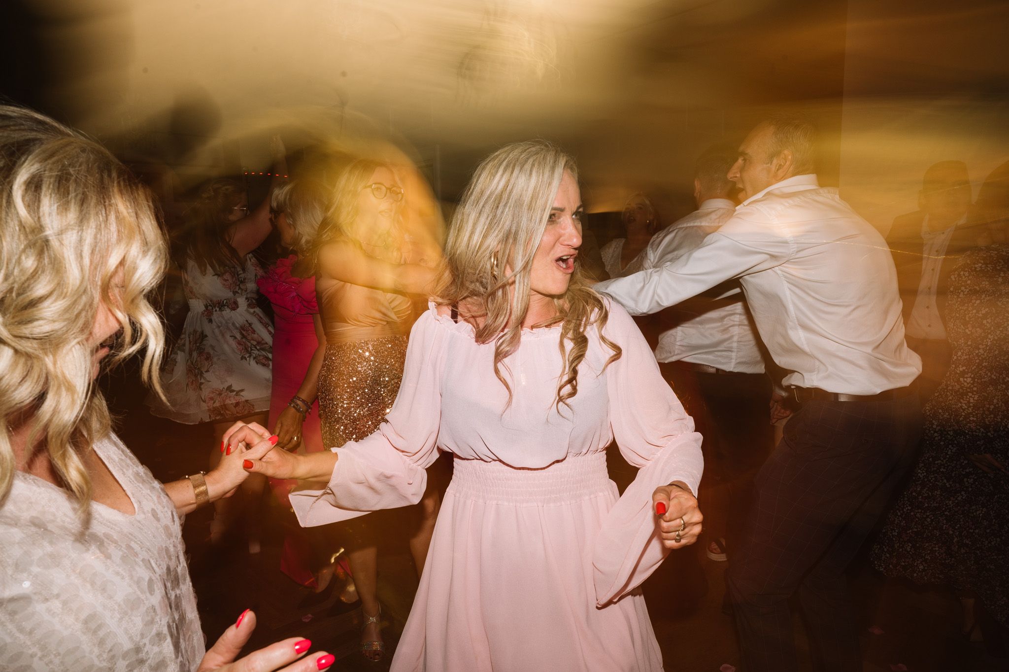 Wedding guests dance and celebrate with motion blur creating dreamy atmosphere under warm lighting.