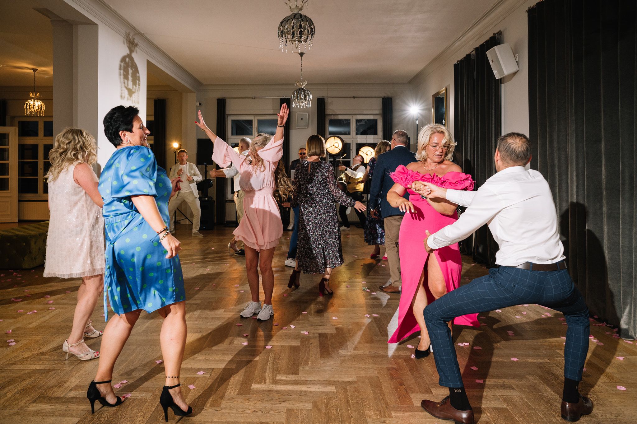 Group of wedding guests dancing on wooden floor in colorful formal attire.