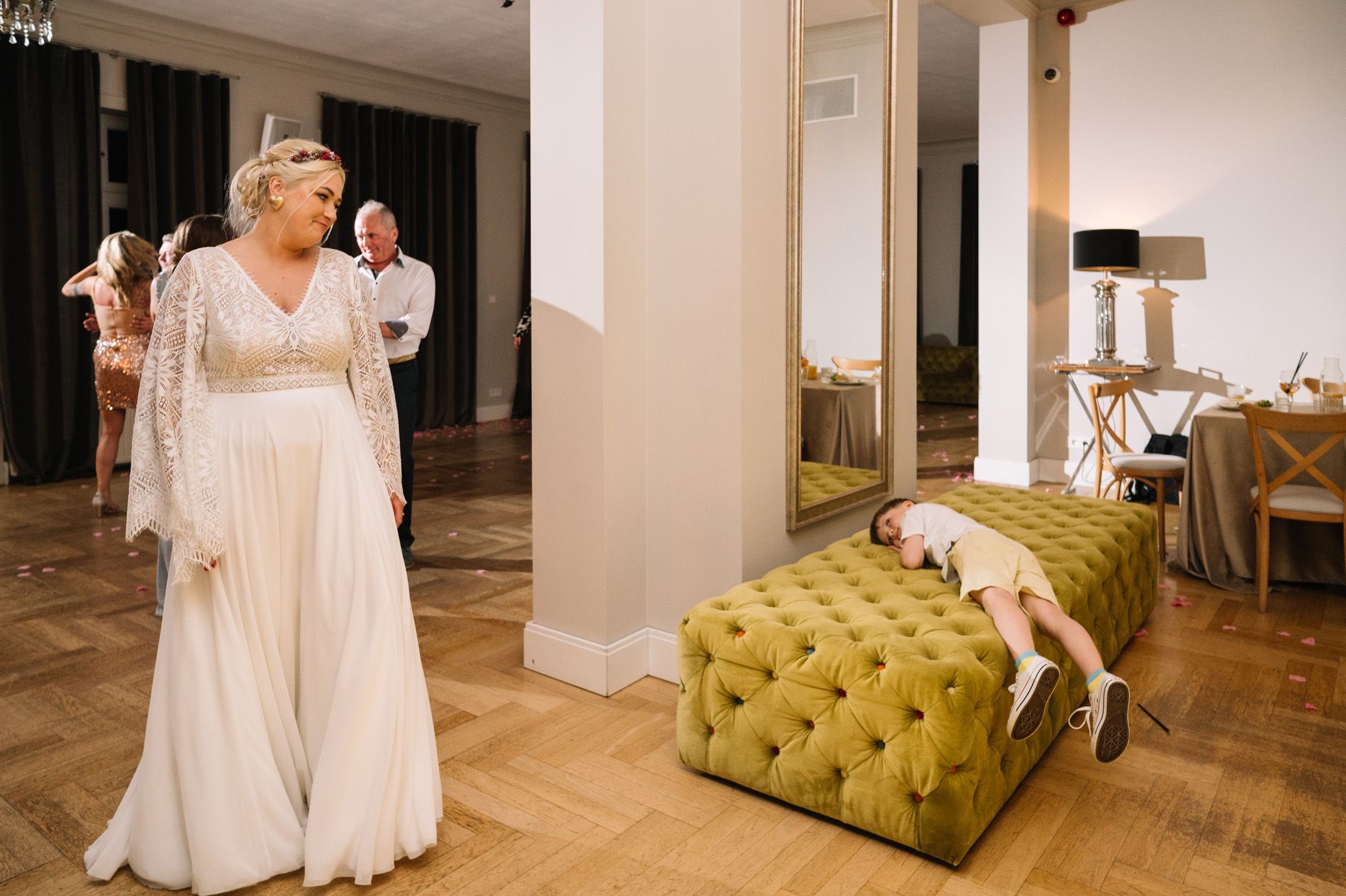 A long white dress flows elegantly as a small dog rests on a yellow tufted ottoman nearby.
