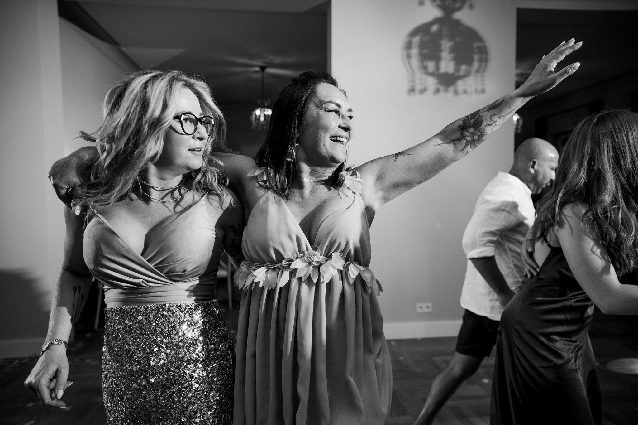 Two women dancing energetically at a party, one wearing a sequined dress and glasses, the other in a flowing gown.