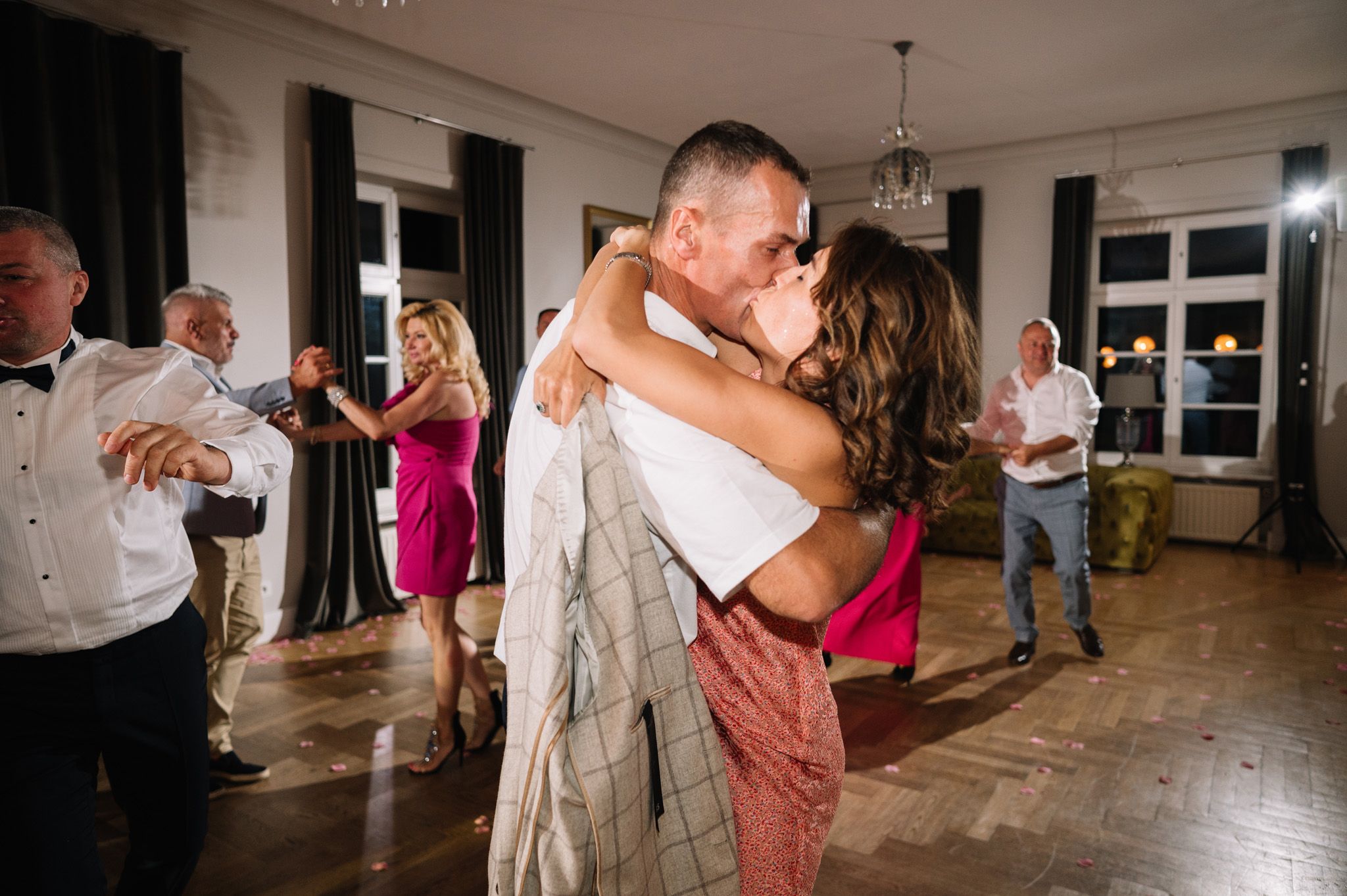 Couples dance together at an evening party or reception in an elegant room with wooden floors and large windows.