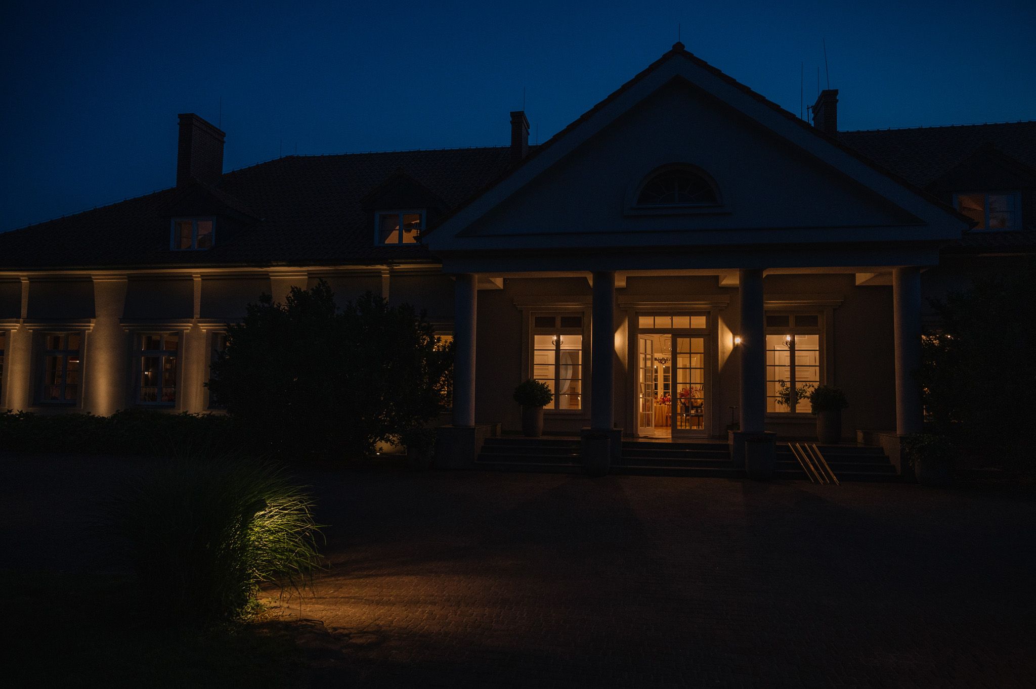 A stately mansion illuminated at night with warm glowing windows and exterior landscape lighting on the facade.