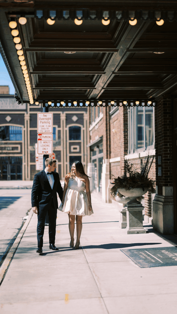 A well-dressed couple walks under a historic building's overhang with decorative lighting and architectural details.