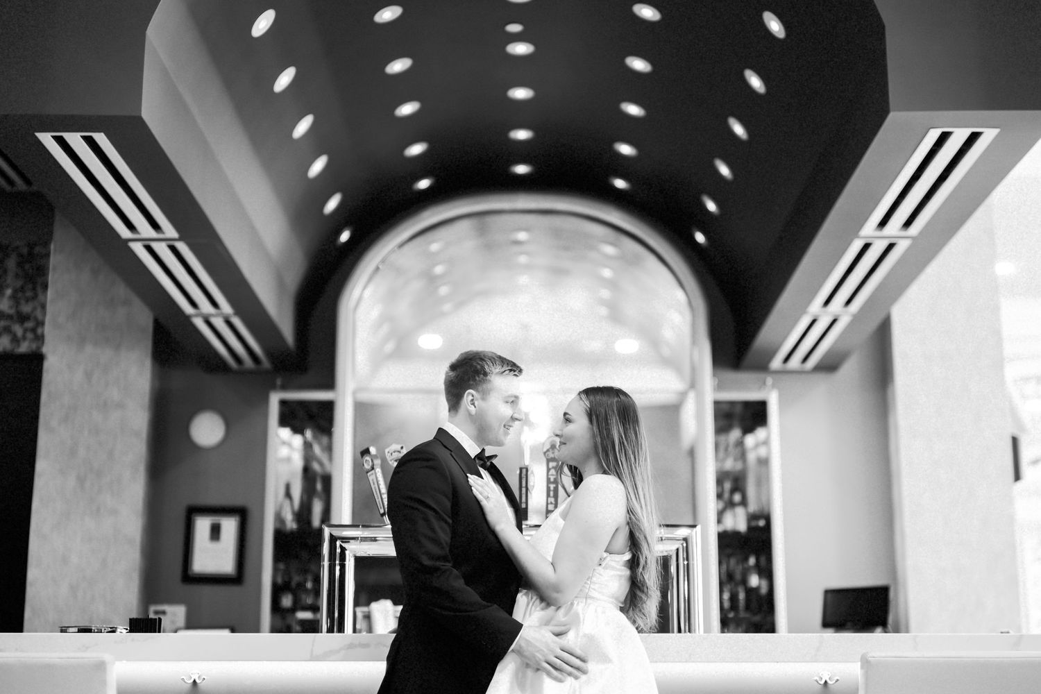 A romantic wedding couple shares a tender moment under an arched entryway with elegant overhead lighting.