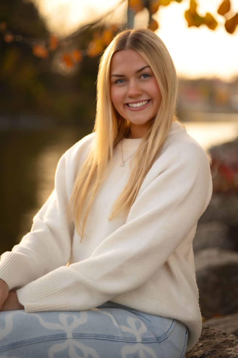 Portrait of someone in a white sweater sitting outdoors during sunset with autumn foliage.