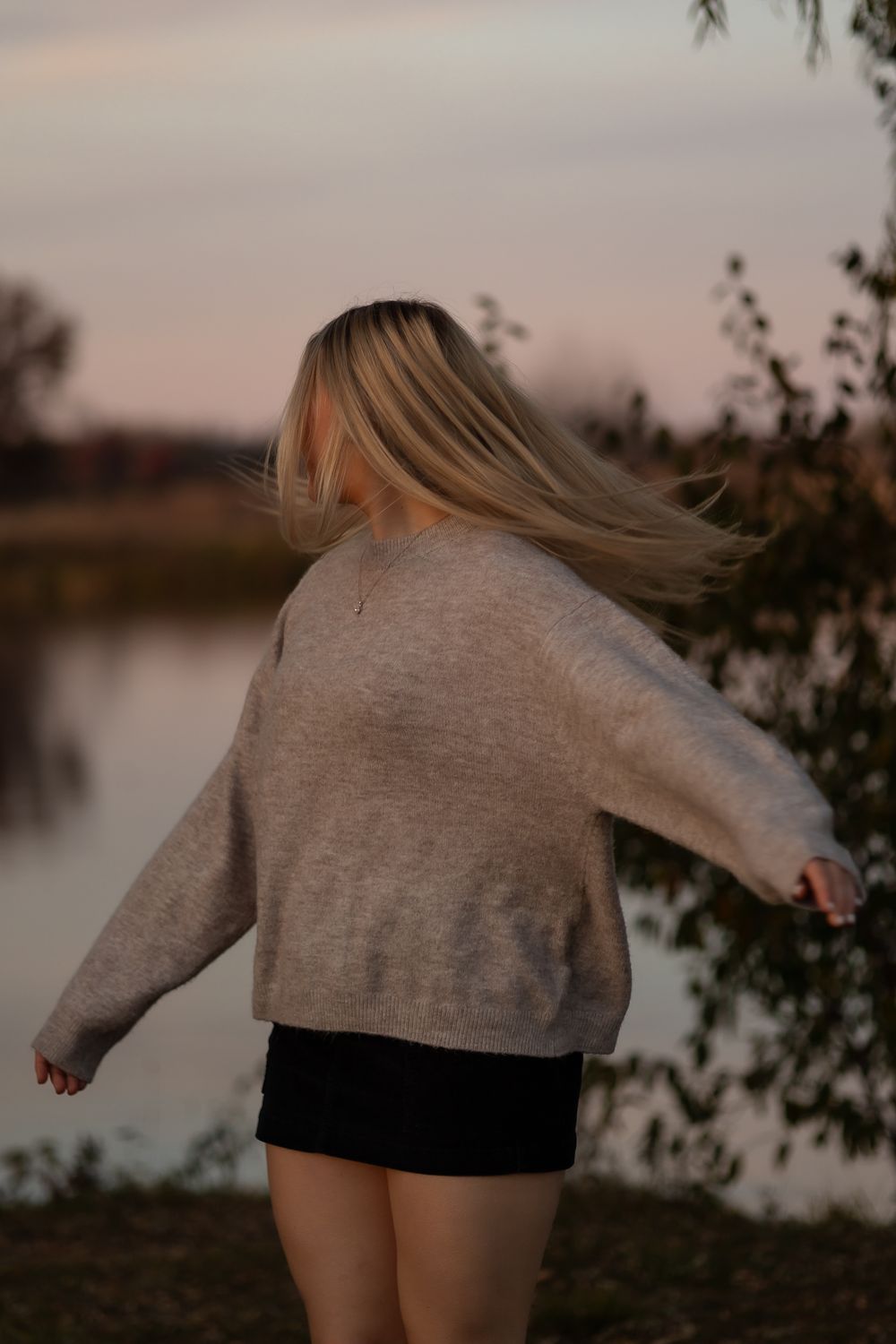 A person in a gray sweater and black shorts spins joyfully by a lake at sunset, creating a dynamic motion sequence.