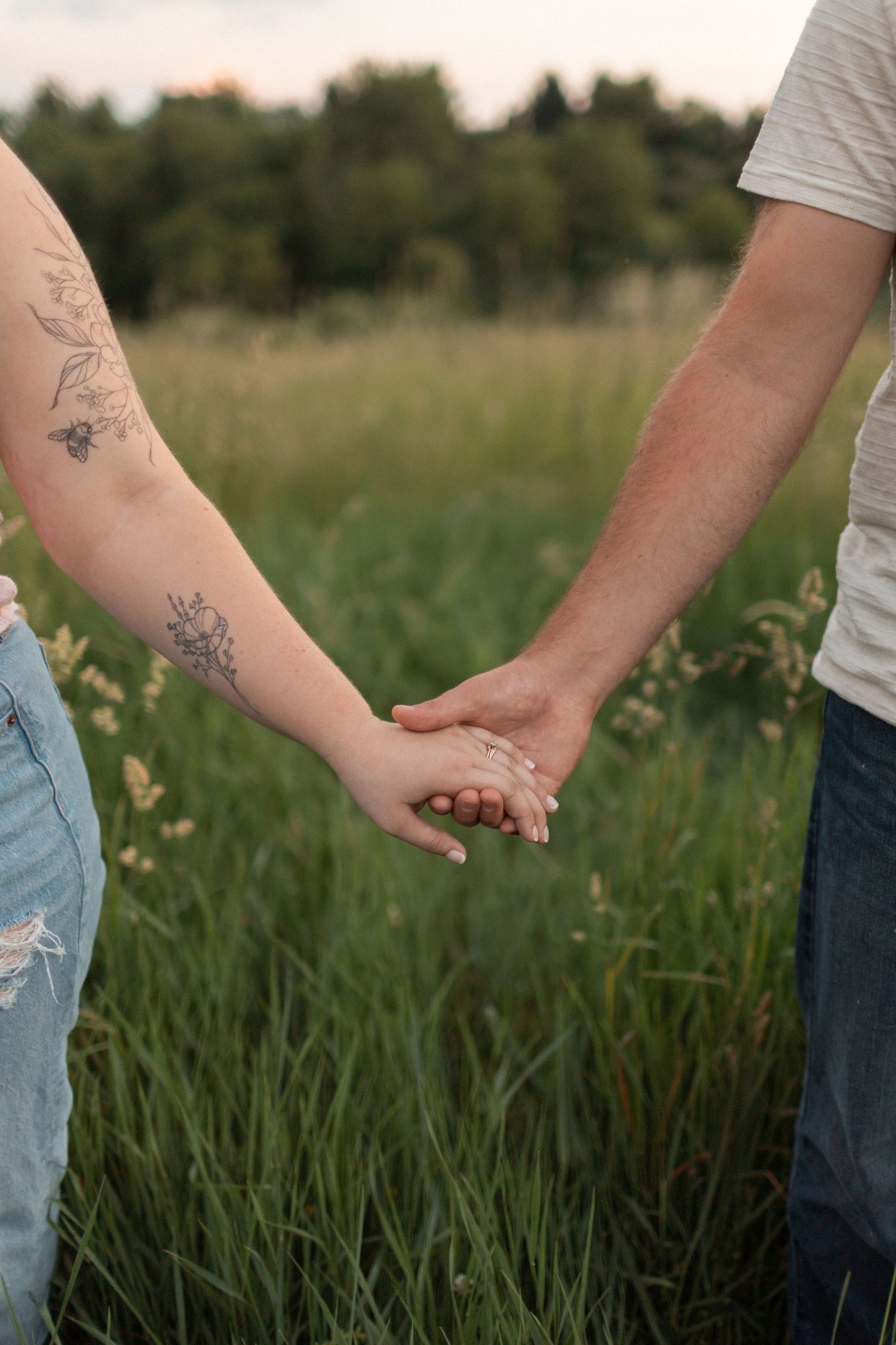 Close-up of couple holding hands in tall grass field showing floral tattoos.