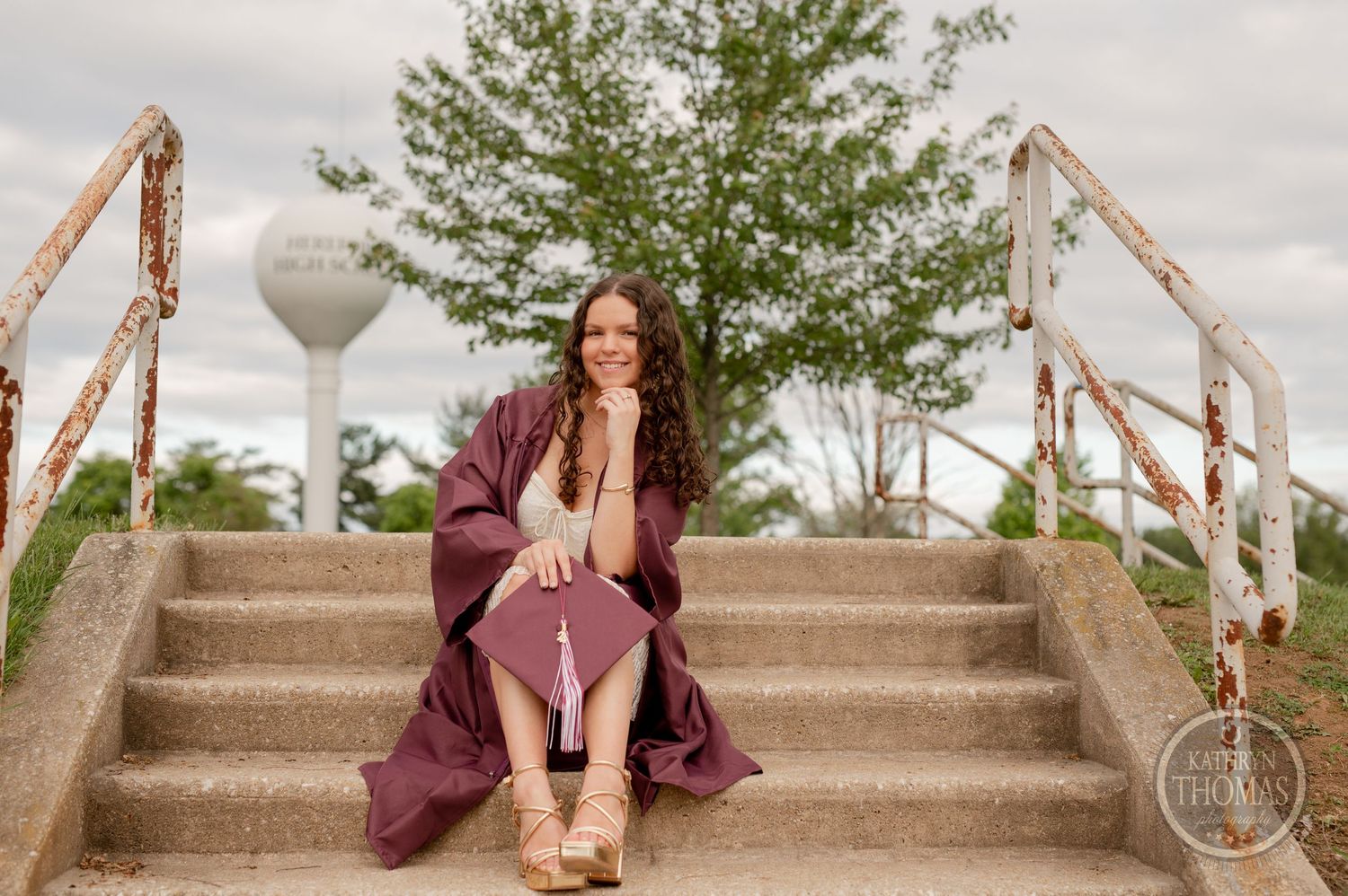 Graduate in maroon gown sits thoughtfully on concrete steps with water tower visible.