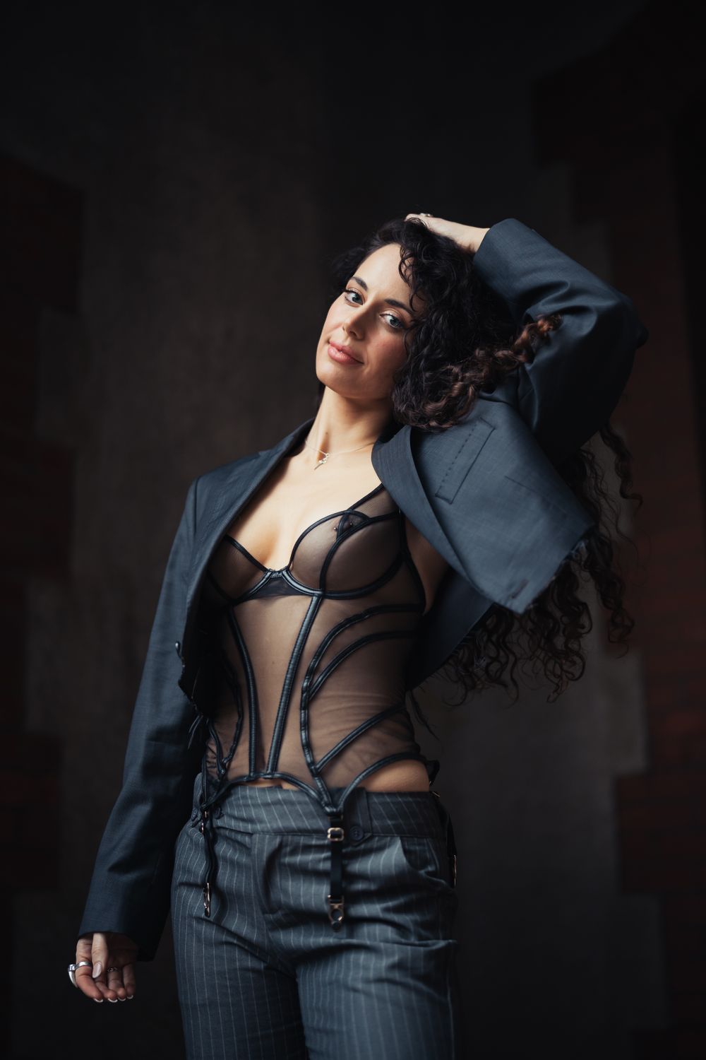 Model poses in pinstripe suit and sheer top against dark studio backdrop.