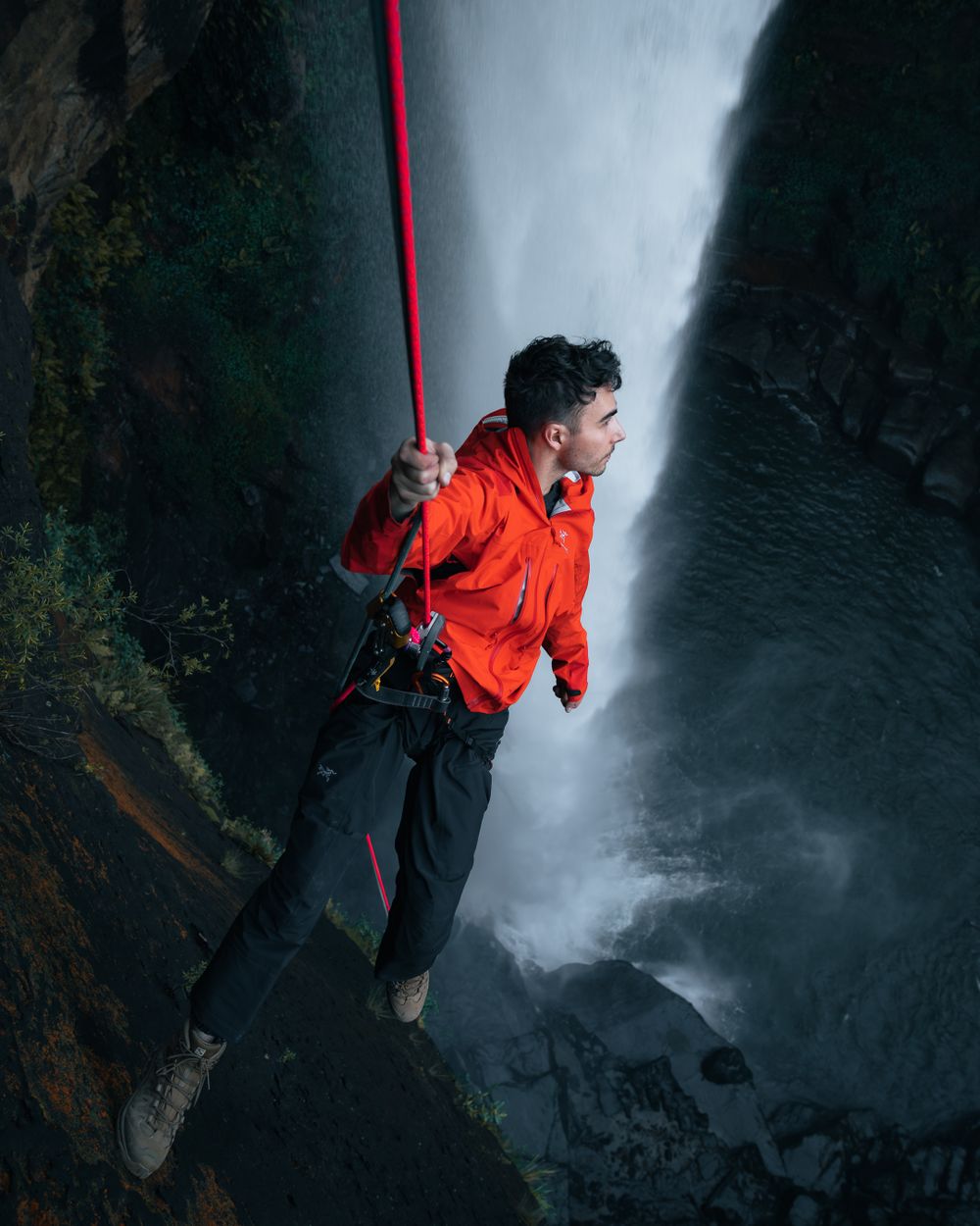 Person in orange jacket rappelling down dramatic waterfall in misty conditions.